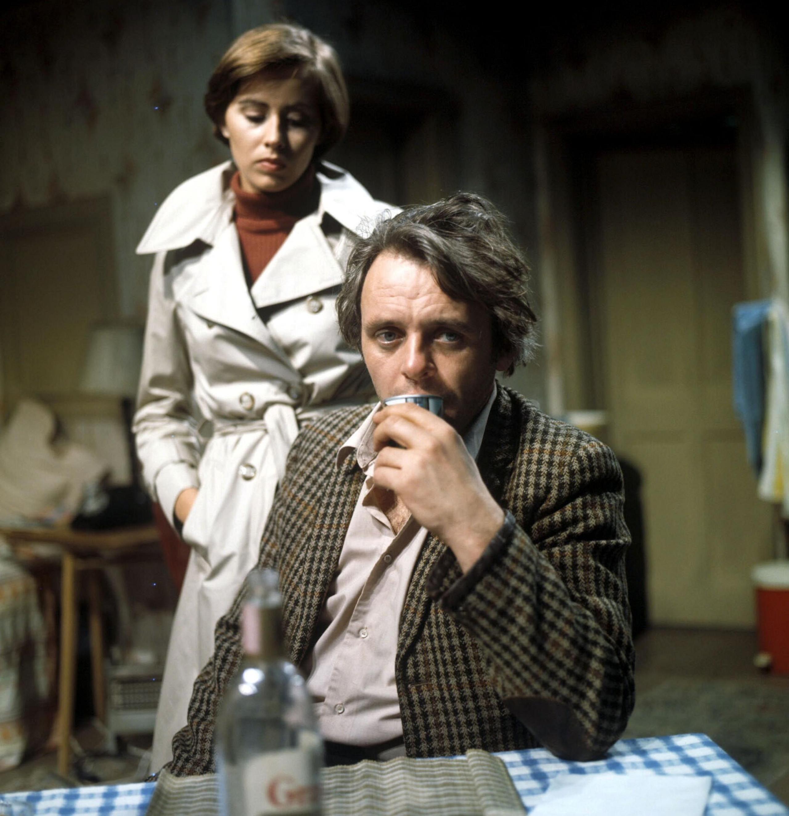 Hopkins looking dishevelled , with checked jacket, shirt with top bottom open, holding a drink, seated at a table with a bottle of alcohol in the foreground.
Kate Nelligan standing in light beige mac, short brown hair, looking sadly at him