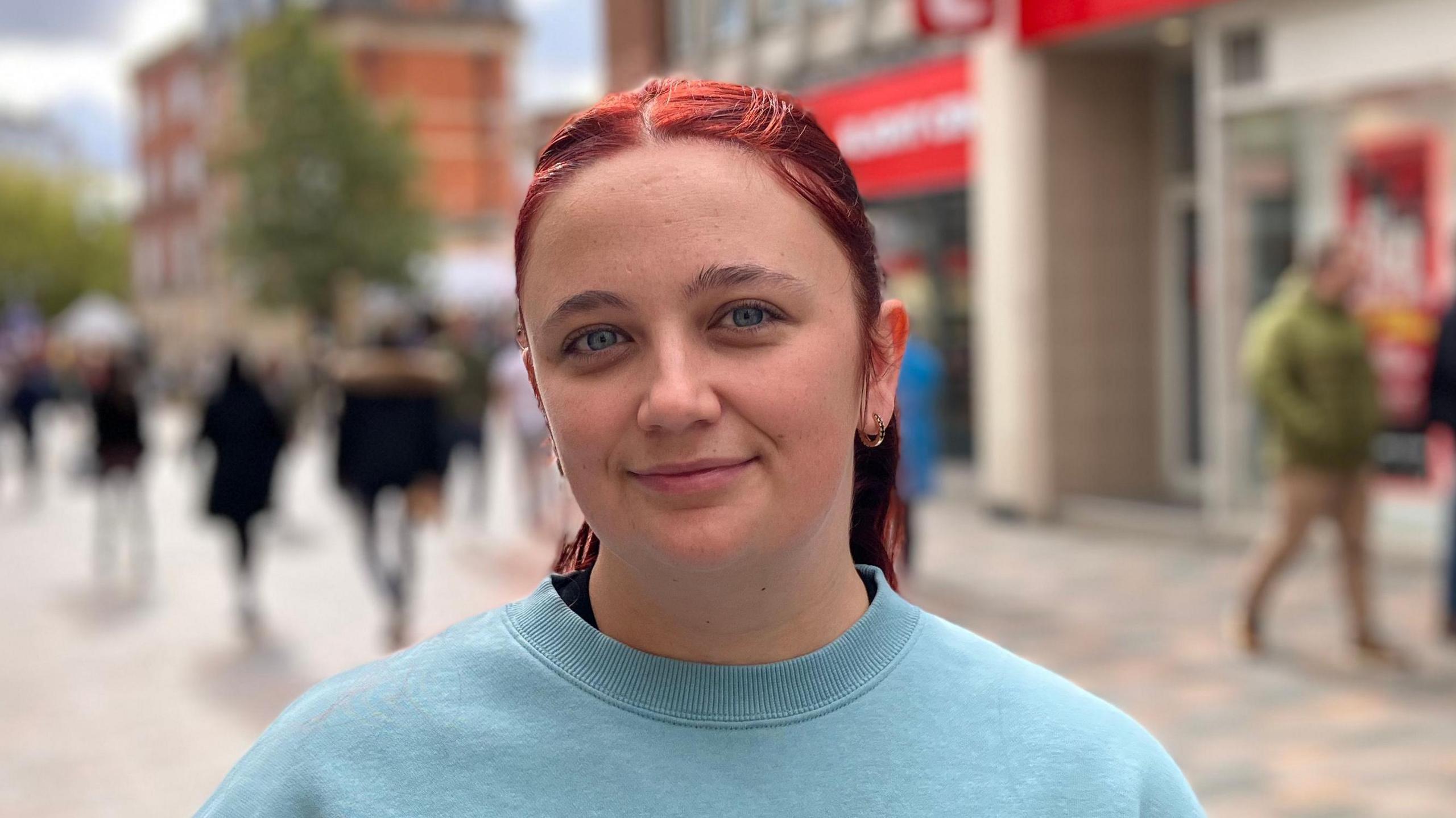 Kerry Smith is wearing a turquoise jumper. She has red hair and is smiling. Behind her are shoppers walking along the High Street.