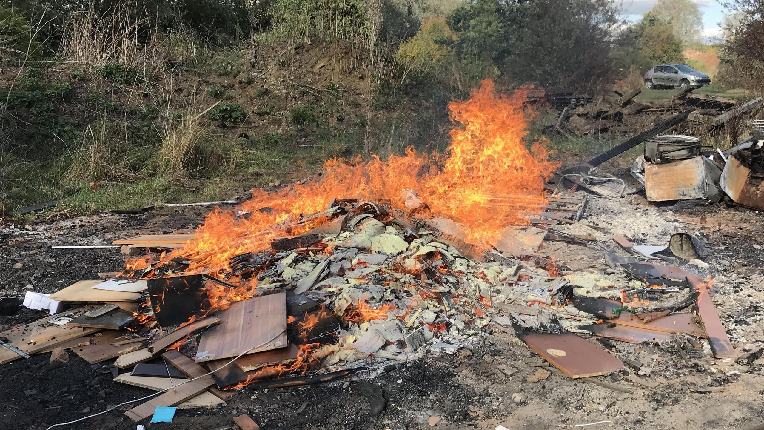 A pile of debris and rubbish is on fire. It is surrounded by green vegetation. A silver car is parked on the horizon.