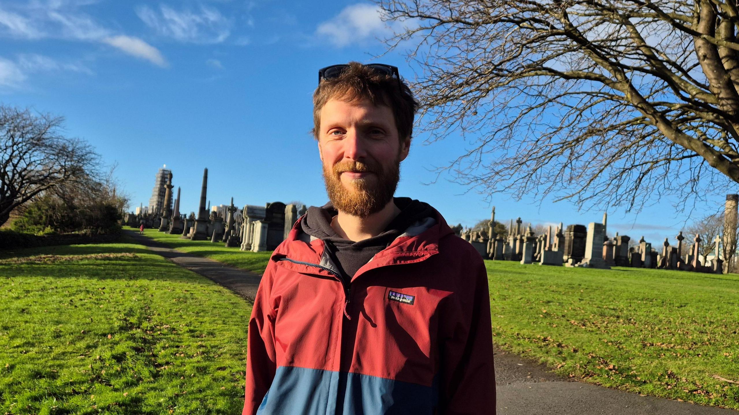 Greg Hawke, who has red hair and a beard, standing outside the graveyard on a sunny day. He is wearing a red and blue jacket, with sunglasses on top of his head. 