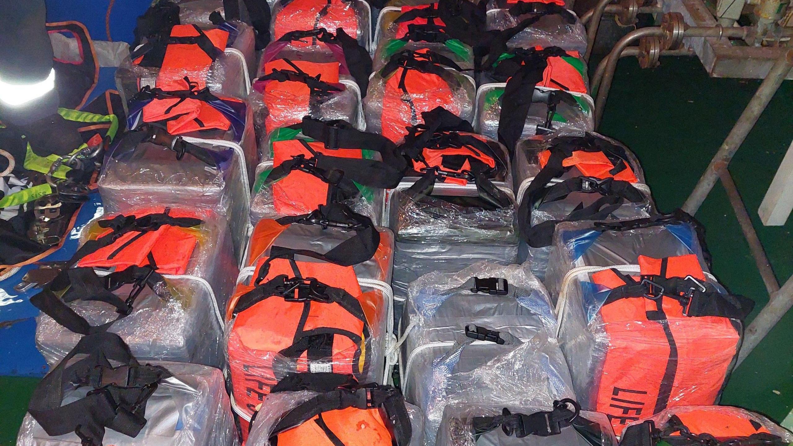 Around twenty large parcels of cocaine, wrapped in waterproof cellophane and covered in orange lifejackets, laid out on the deck of a ship.