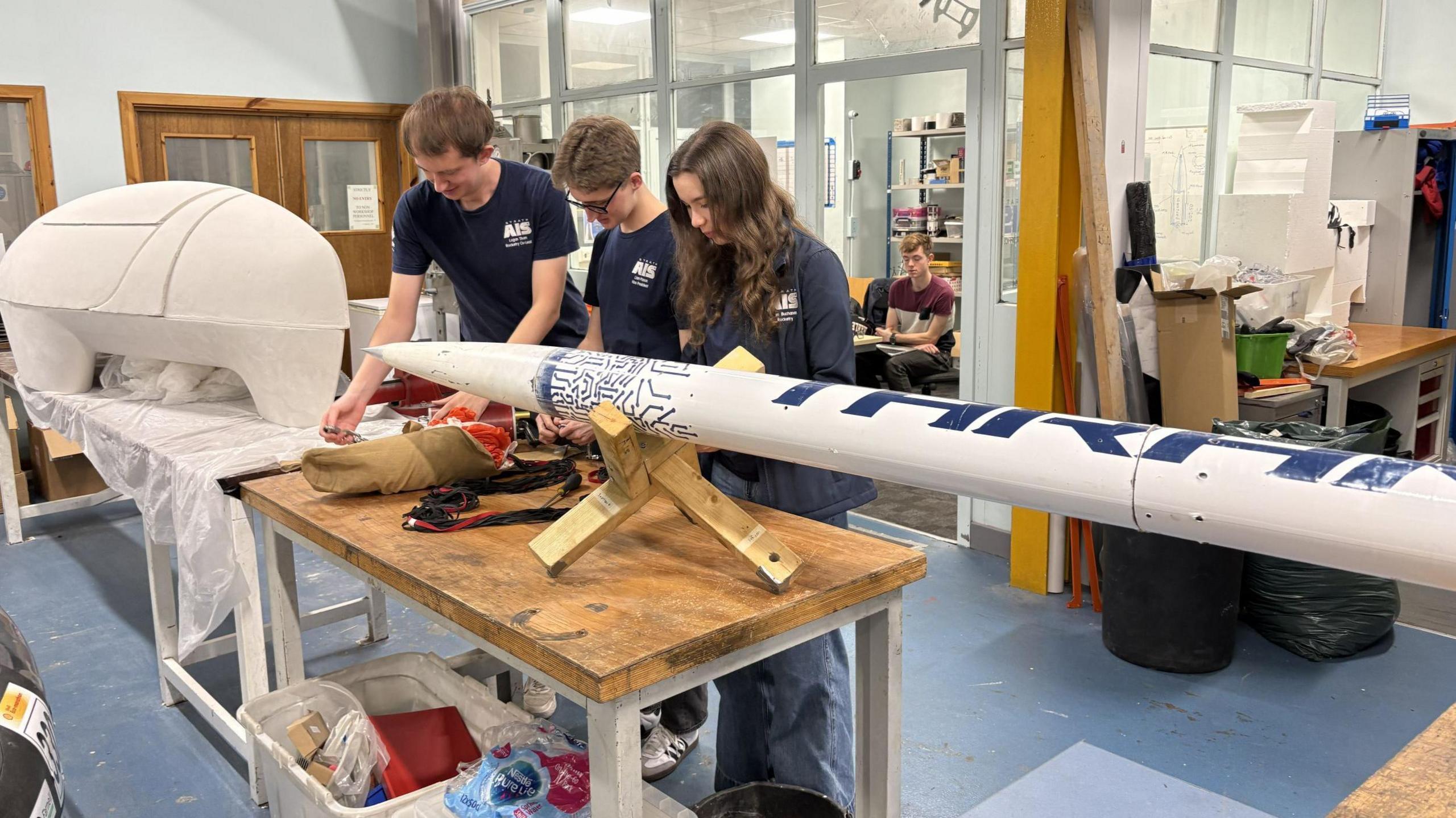 Three students - two males and a female - look over a small rocket on the table in front of them. The rocket sits on a wooden stand as they work on smaller components in the engineering lab.