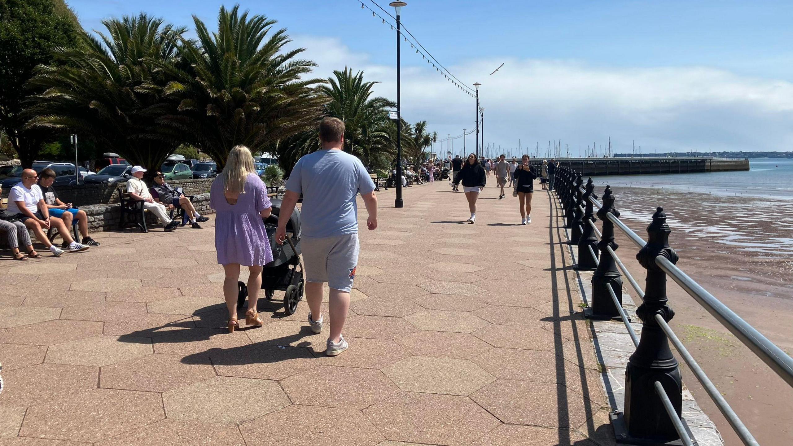 People walking along the seafront. There are people sat on benches on the left, and trees behind them. There are two people walking with a pram at the centre, and the beach is on the right.