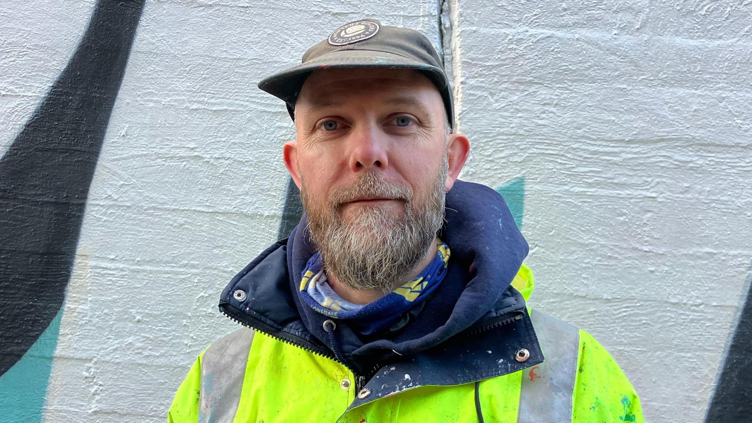 A man with a brown cap, brown and grey beard, and large neon jacket looks at the camera with a neutral expression. The painted white, black and blue wall is just behind him.