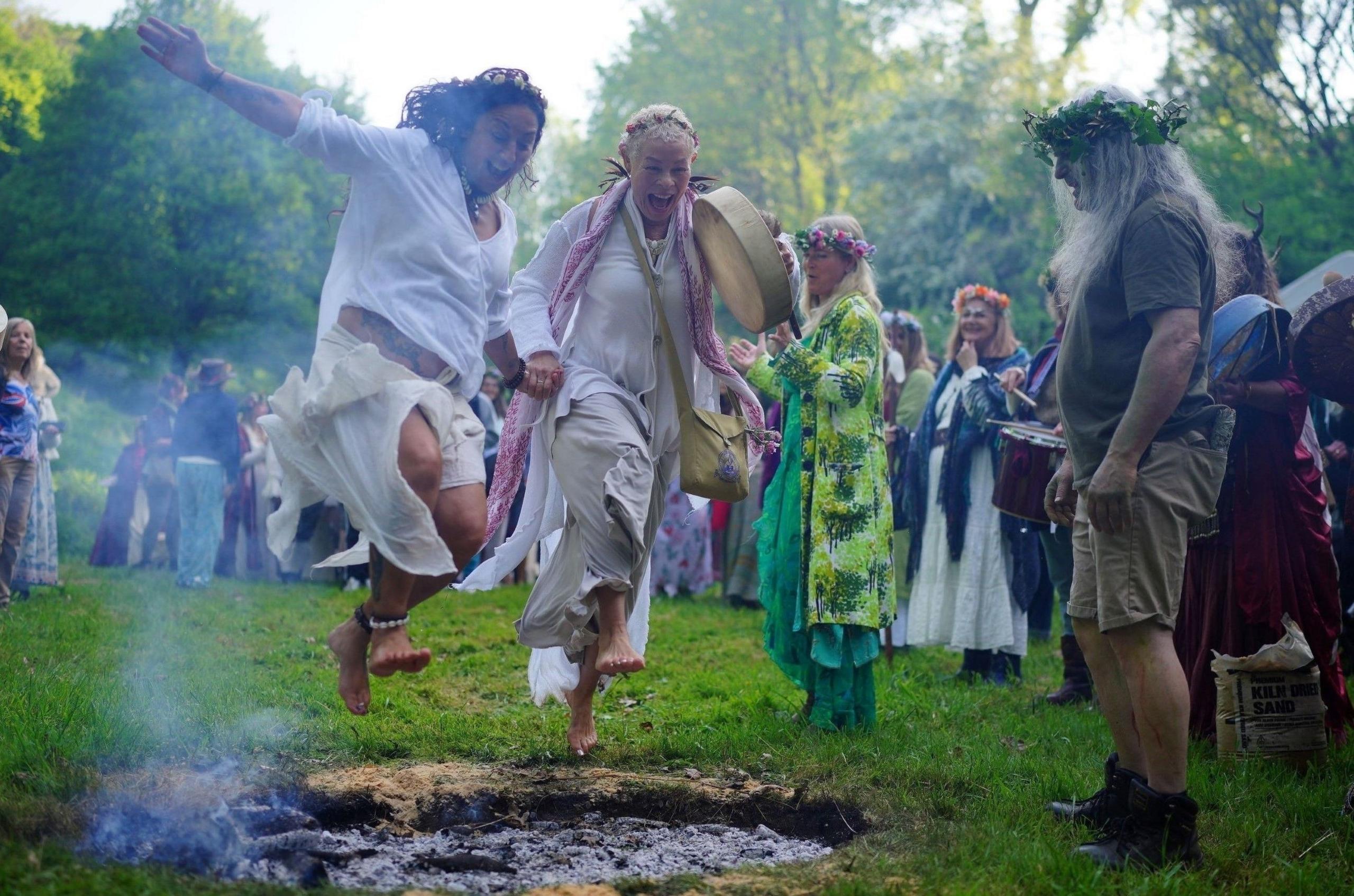 Two women in traditional white pagan outfits jump over a smoking fire while watched by other people during a Beltane ceremony in Glastonbury