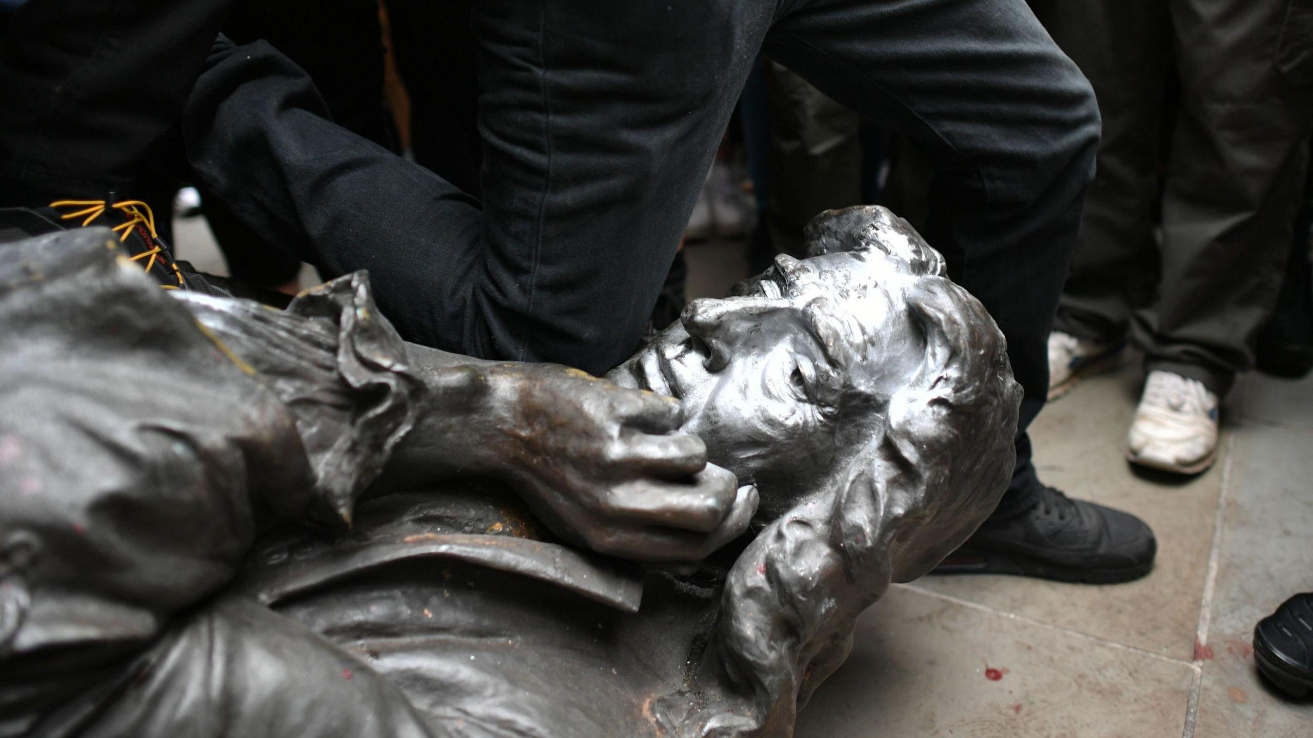 The Edward Colston statue on the floor after being toppled by protesters in Bristol. The metal figure depicts Colston with his chin resting in his hand. A protester wearing black jeans is kneeling on the statue's neck.