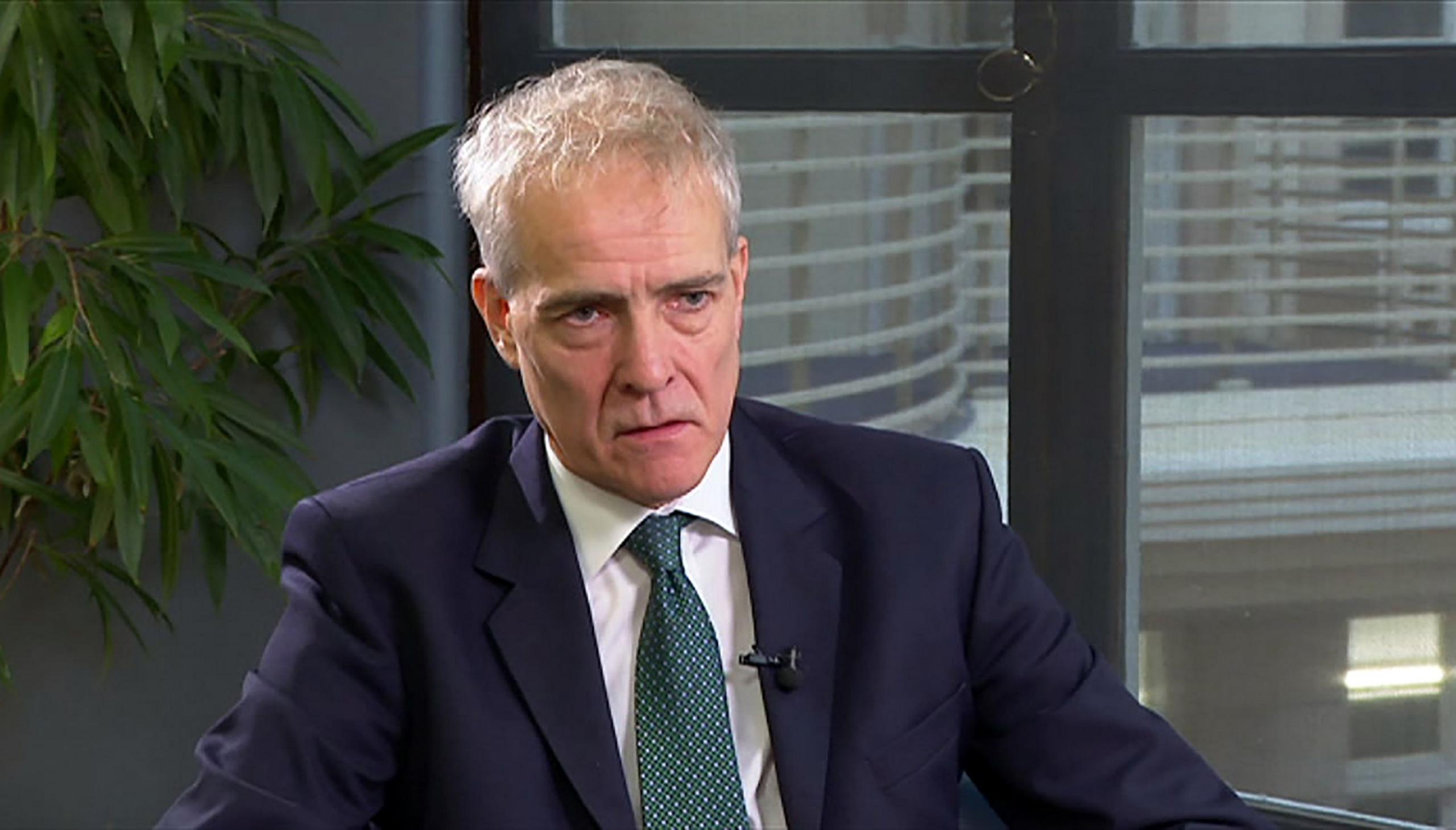 Charlie Taylor wears a white shirt, green tie and dark blue suit with a clip mic attached to the lapel. He is sitting near a window and looking off to the right