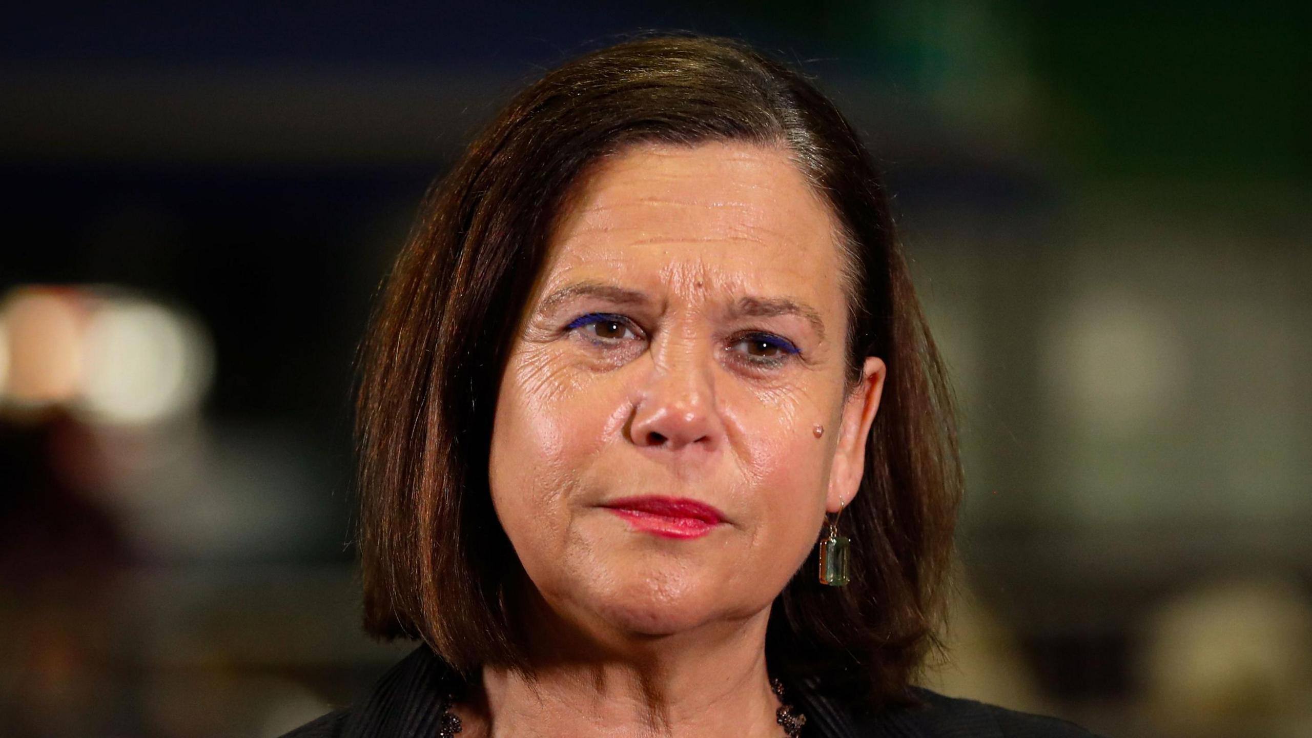 A woman with dark brown hair cut into a bob looks off to the side of the camera. She has brown eyes and is wearing red lipstick. You can see the shoulders of a black formal jacket and a dark metal necklace. The background is blurred. 