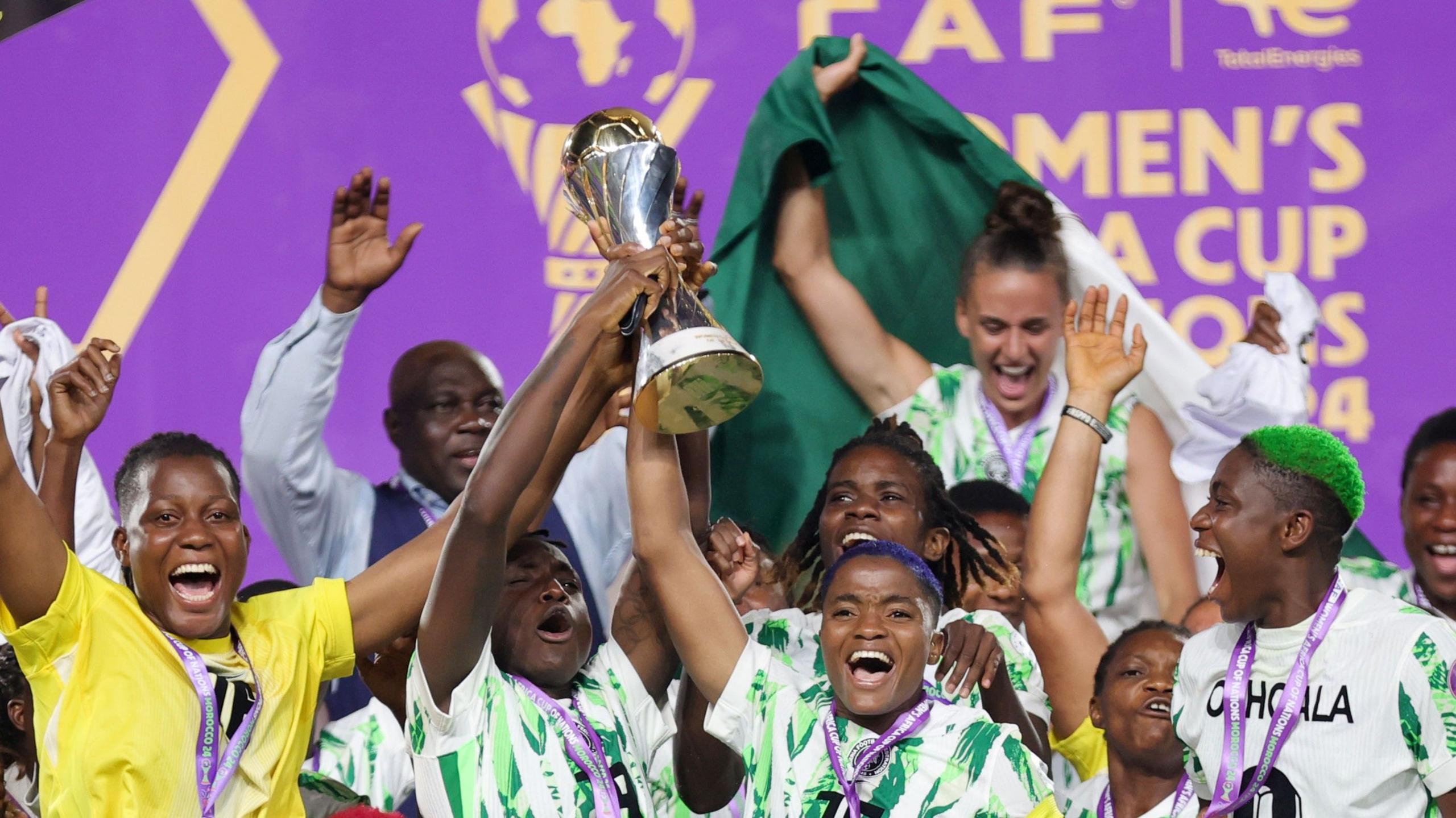 Nigeria's women's football team on the pitch lifting the 2024 Women's Africa cup of Nations trophy. The outfield players are in green and white while the goalkeeper Chiamaka Nnadozie in the bottom left wears yellow. The players all have medals around their necks on purple ribbons
