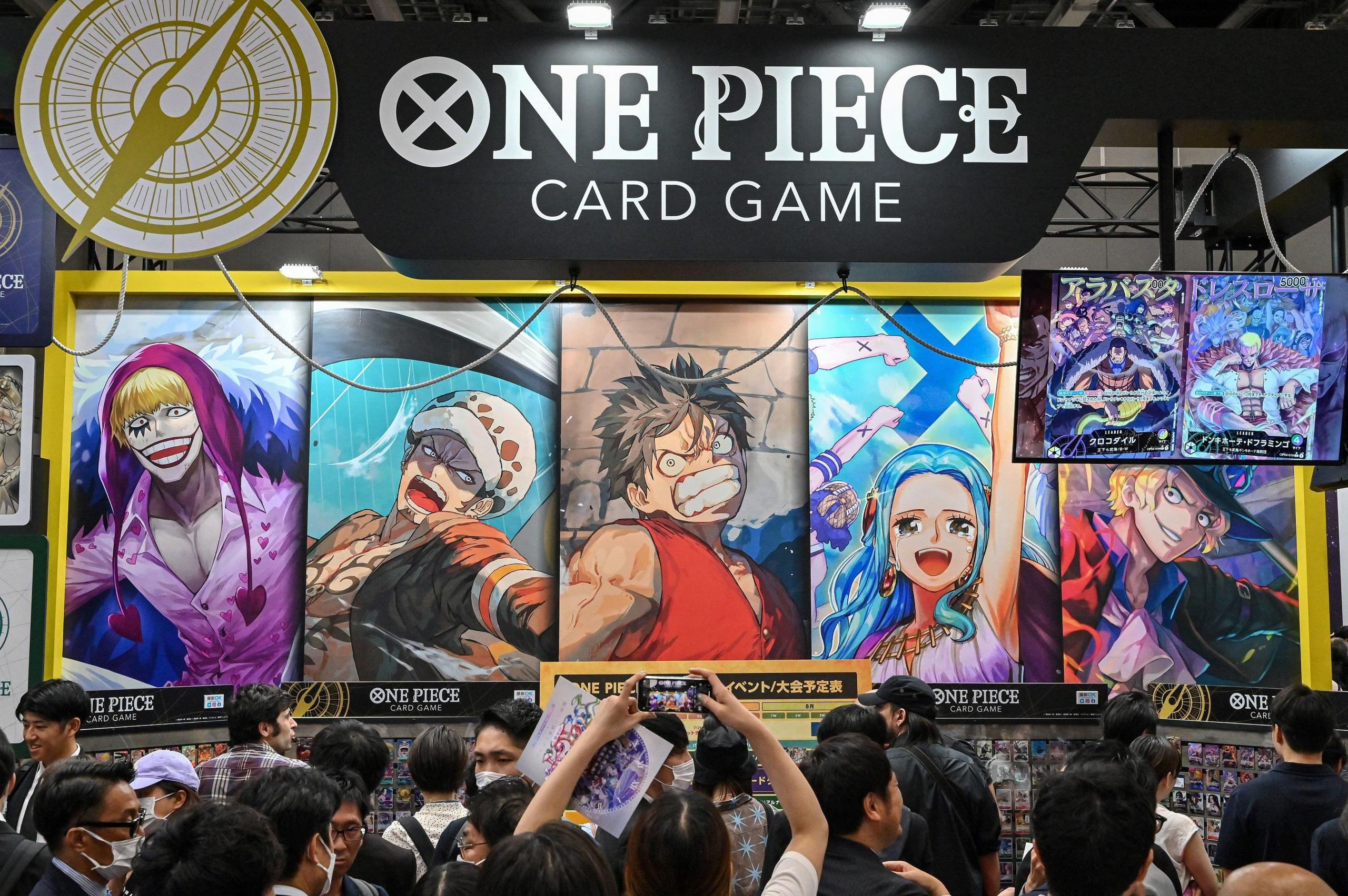 A crowd gathers in from of a giant One Piece card display at a convention in Japan. The cards have various characters drawn in the "manga" style. A banner sits above them with the One Piece logo.