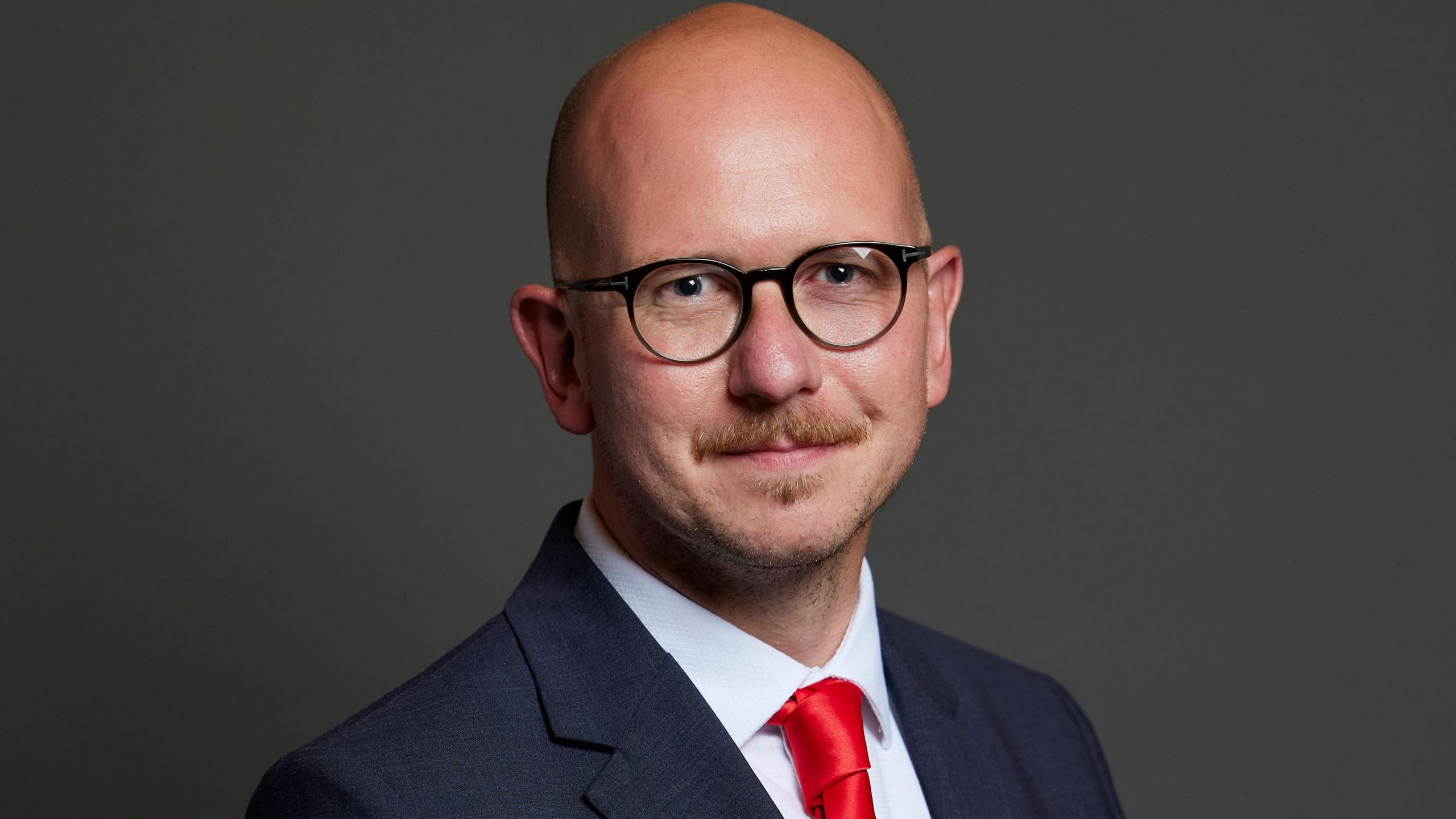 Brian Leishman, a bald man with glasses and moustache is wearing a blue suit, white shirt and red tie. He is smiling, turned to his right.