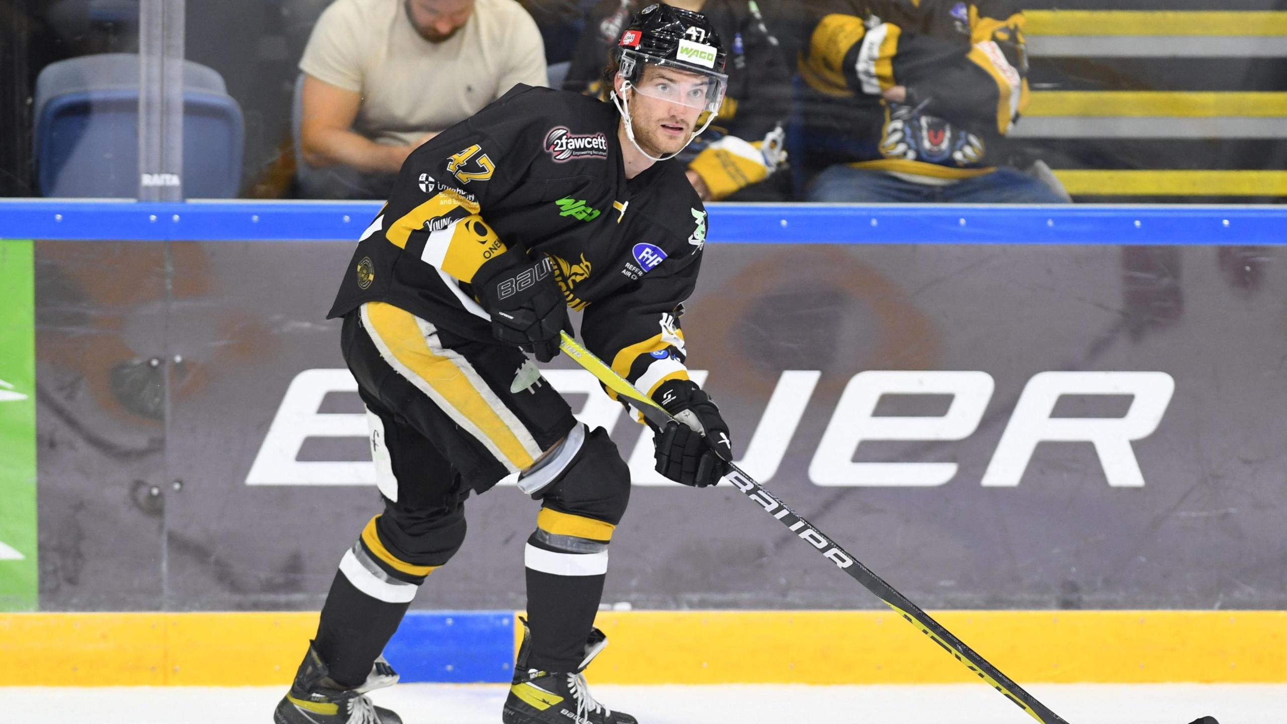 Adam Johnson on the ice wearing a black helmet and jersey, black and yellow shorts, and black socks and skates