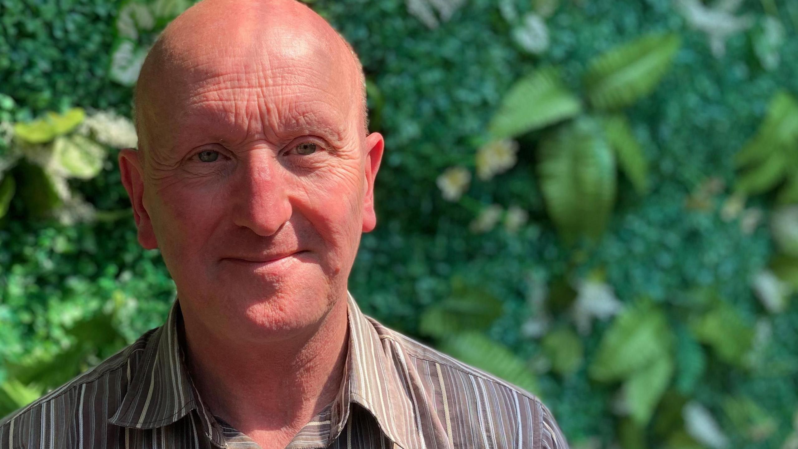 Man with brown and white striped shirt smiling and looking at the camera. A hedge or bush can be seen behind him.