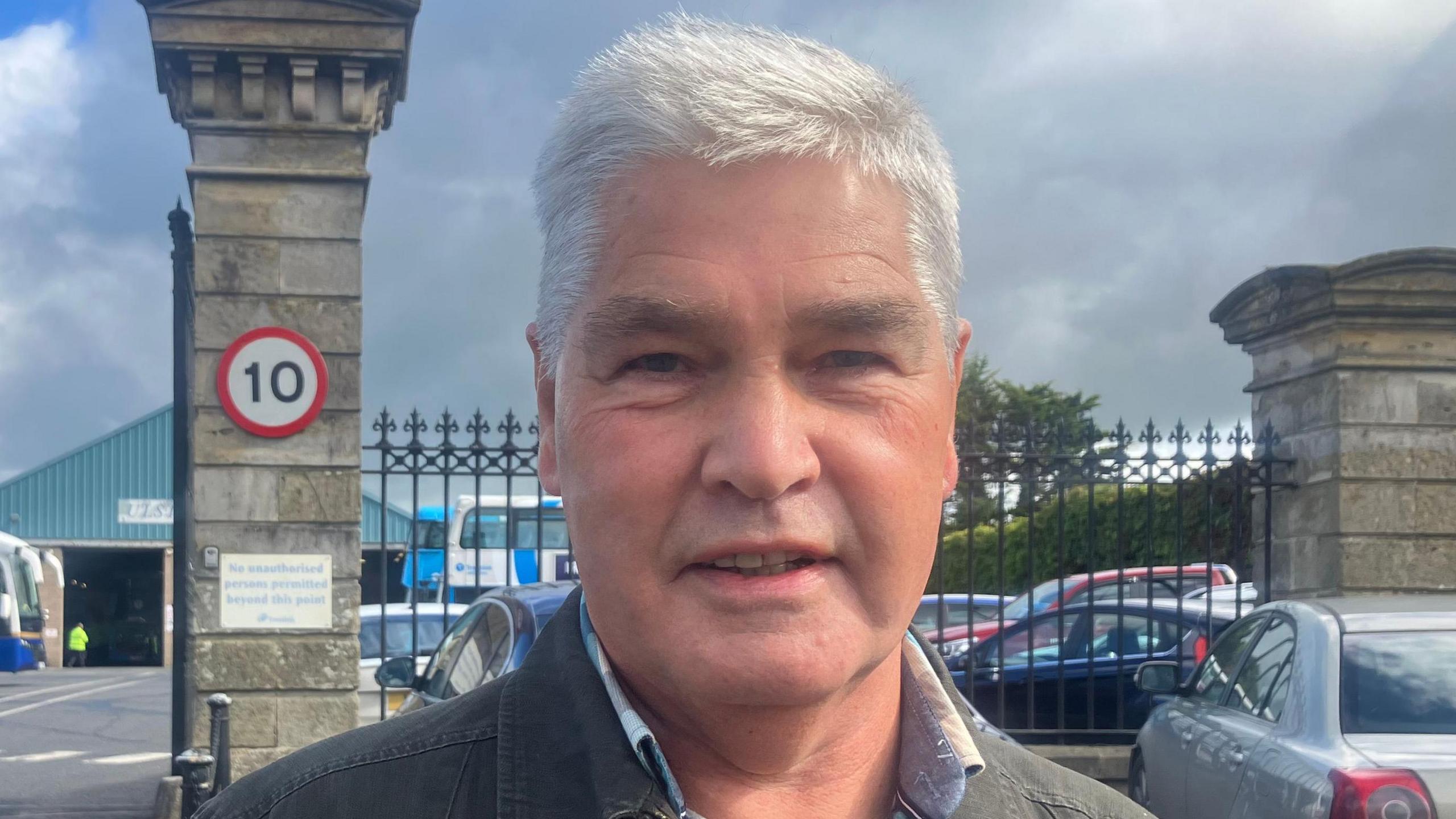 William is standing in front of a bus station. There are buses and cars in the background. He has white hair and wearing a grey jacket with a blue and white shirt.