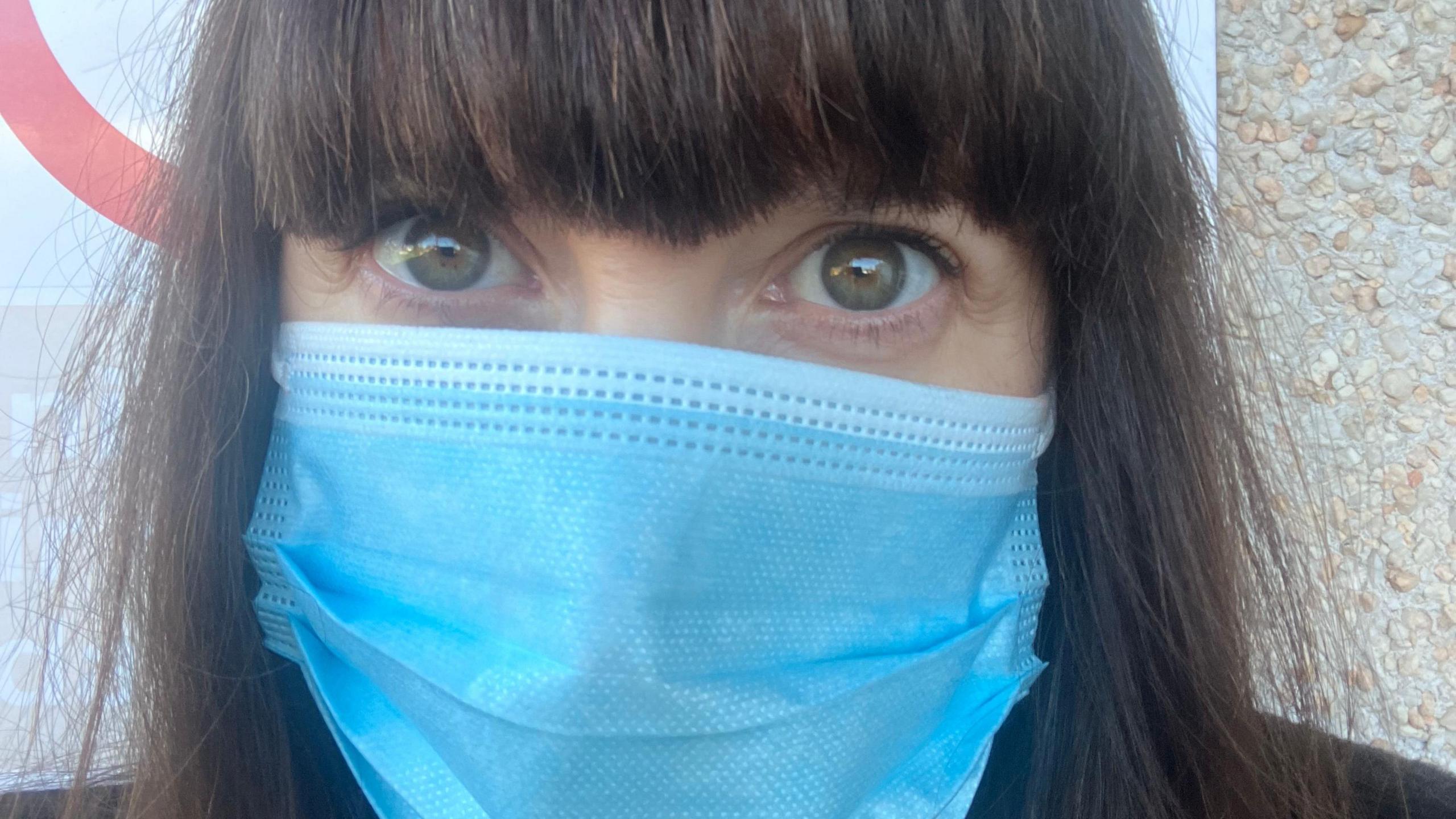 A close up selfie of journalist Nicola Bryan's face. She is wearing a blue face mask and has long brown hair. 