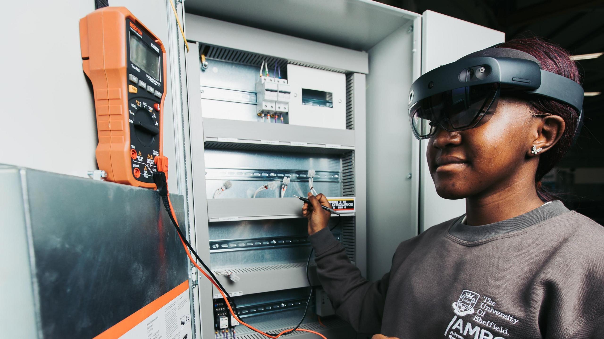 A young woman wearing safety goggles and wearing a green jumper with an Advance Manufacturing Research Centre logo performs tests on some electrical equipment