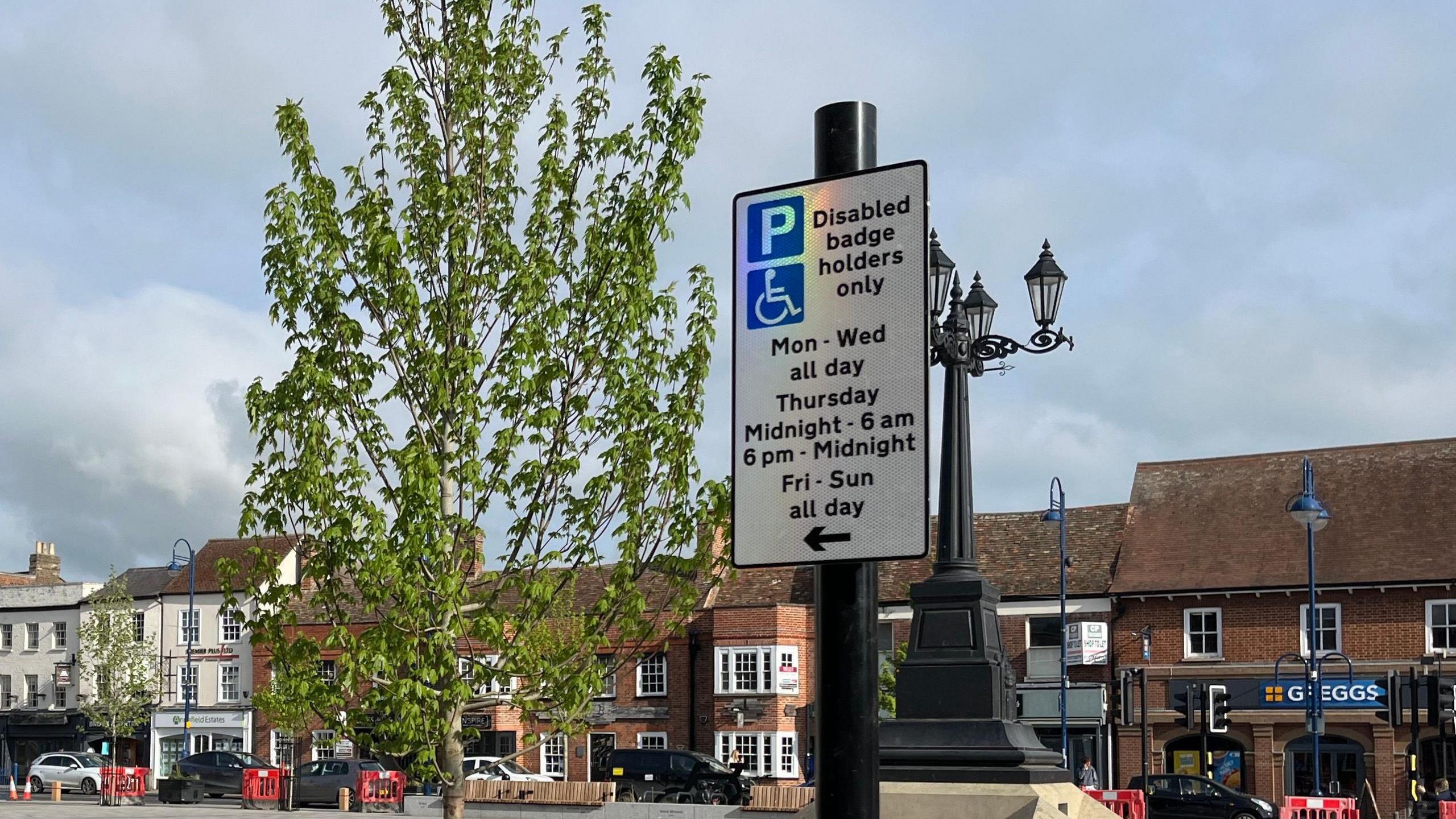 A sign on St Neots Market Square signposting a disabled parking space. It reads 'disabled badge holders only'. In the background is the high street with a Greggs in view.