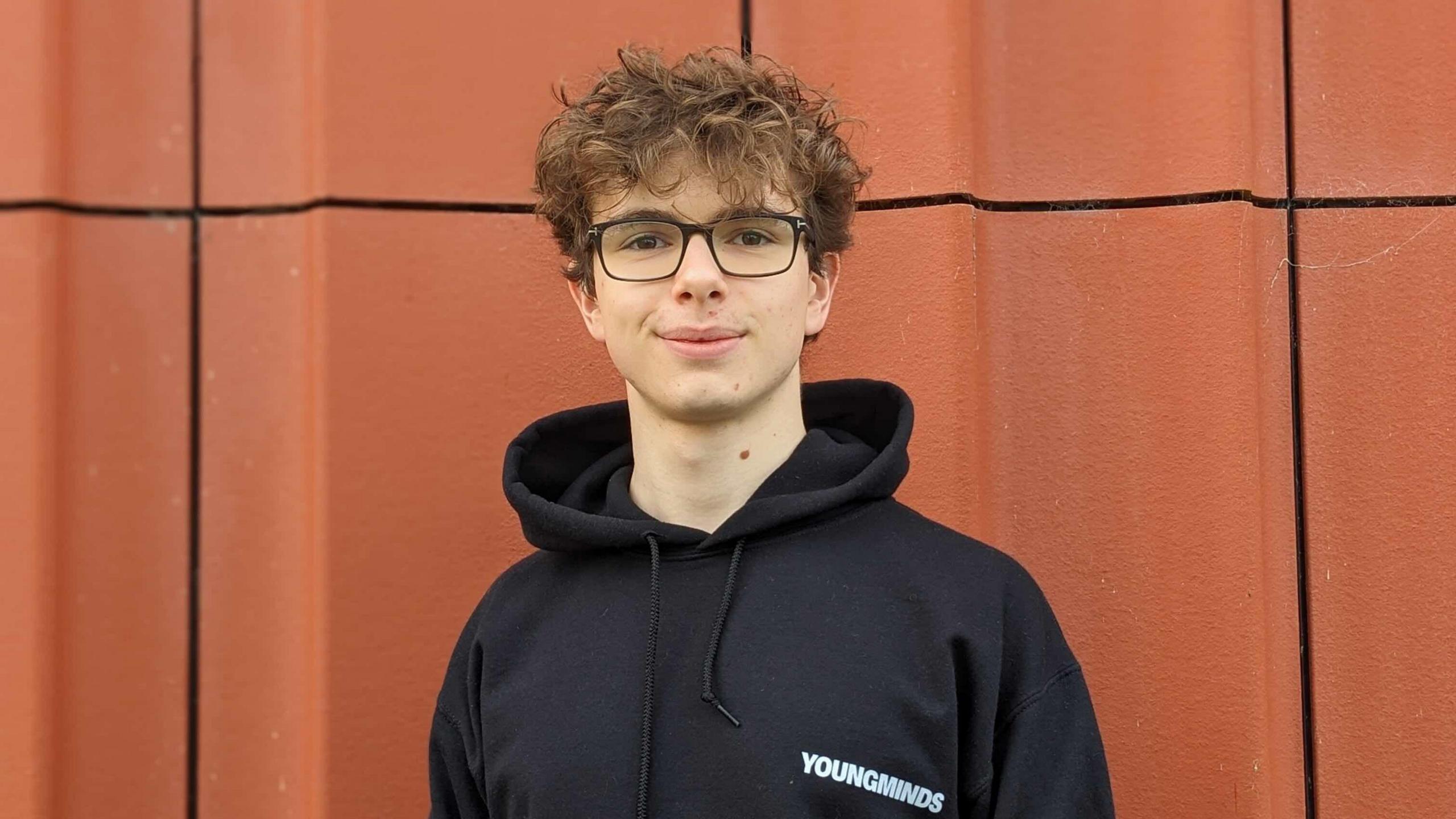 Stefano pictured in front of a burnt orange textured wall. He has short brown hair, black glasses and is wearing a black hoody and smiling at the camera.