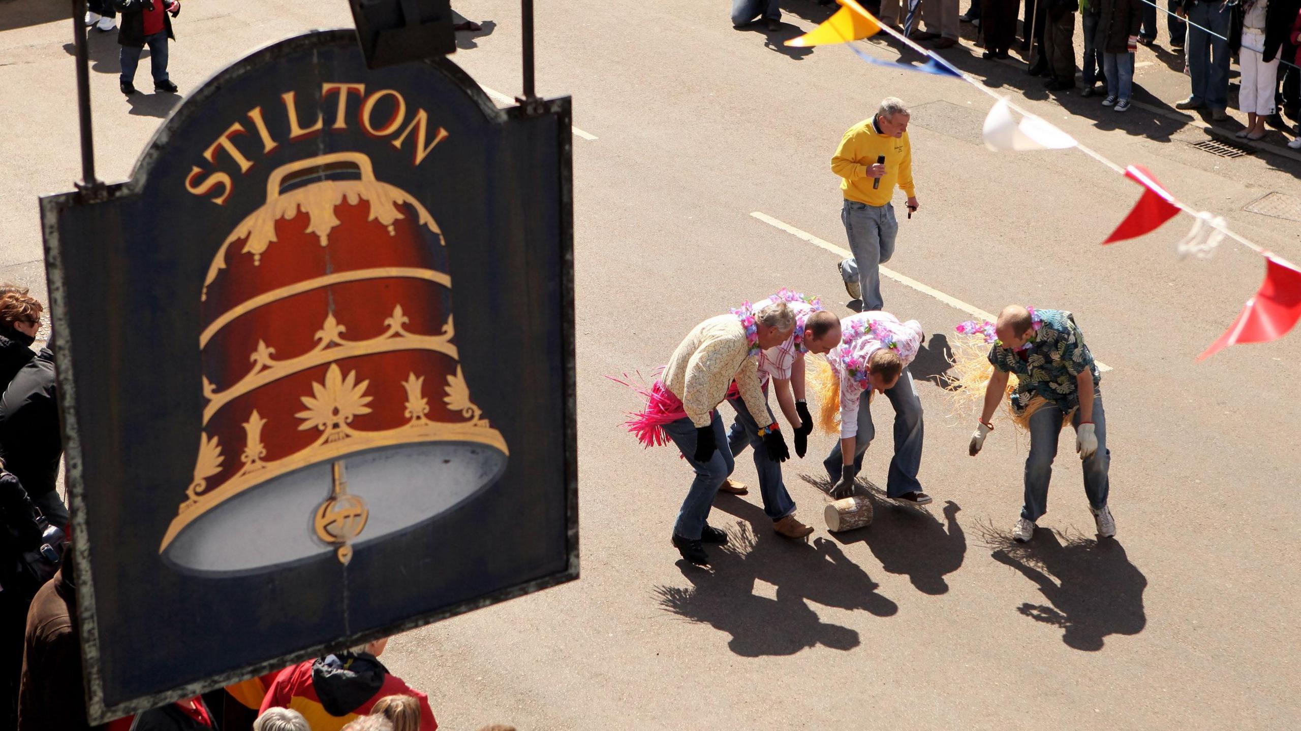 An aerial view of four people, who are dressed in fancy dress, rolling a cheese prop down a road. On the left is a black pub sign that has a red and gold bell on it, with Stilton written in gold writing.