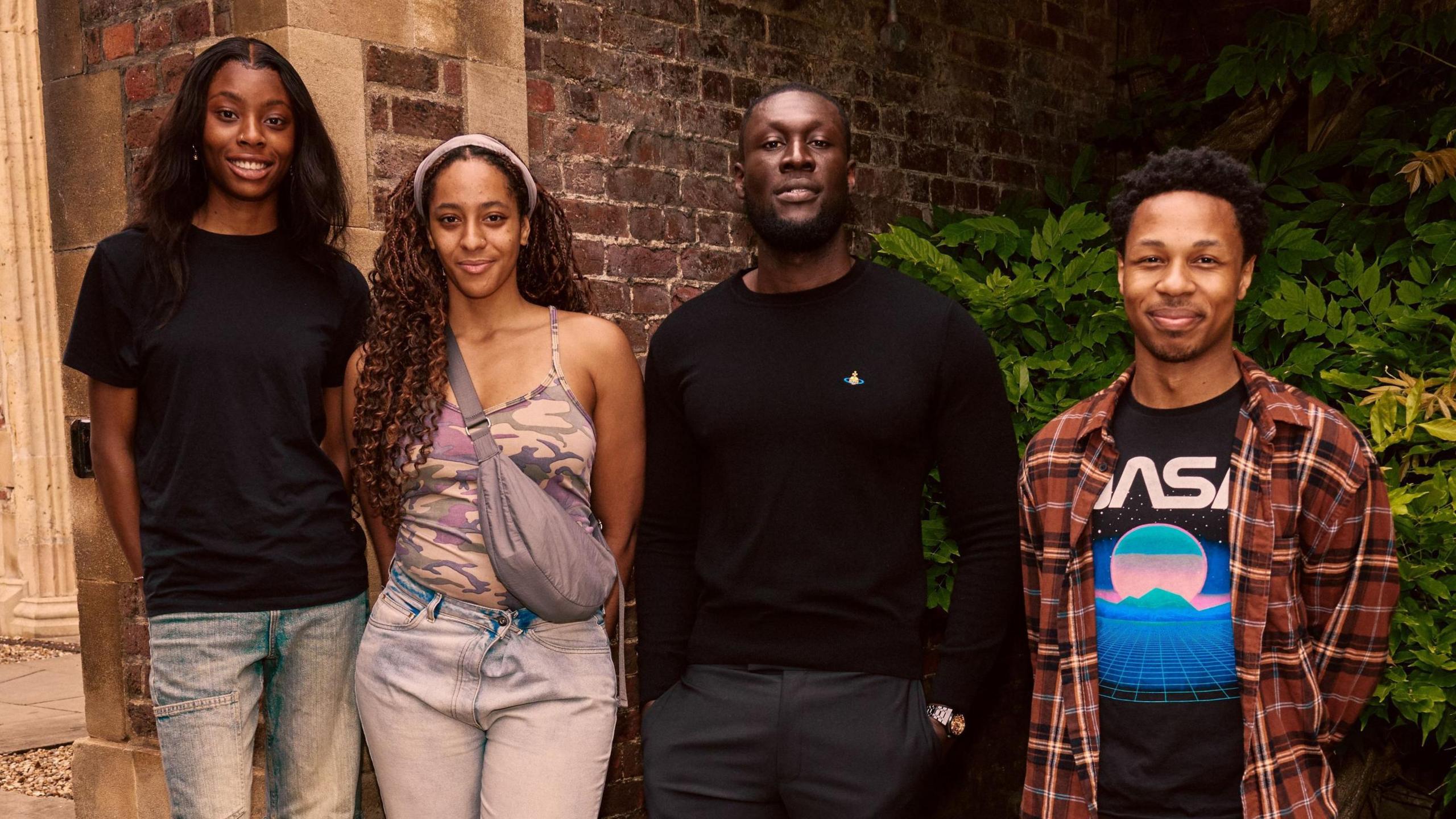 The rapper Stormzy with student Destiny, Drew and Dimario alongside him. They stand outside a brick building with shrubbery.