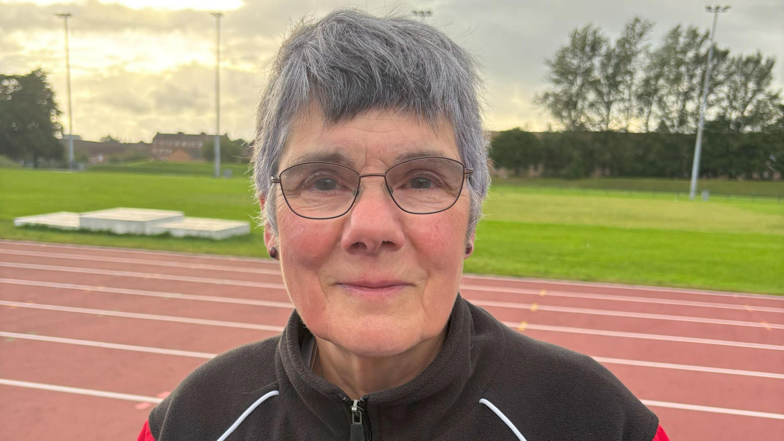 Athletics coach Caroline Johnston - a woman with grey hair and glasses - standing on a running track, with grass behind her and houses beyond that. She is wearing a black tracksuit with white and red trim.