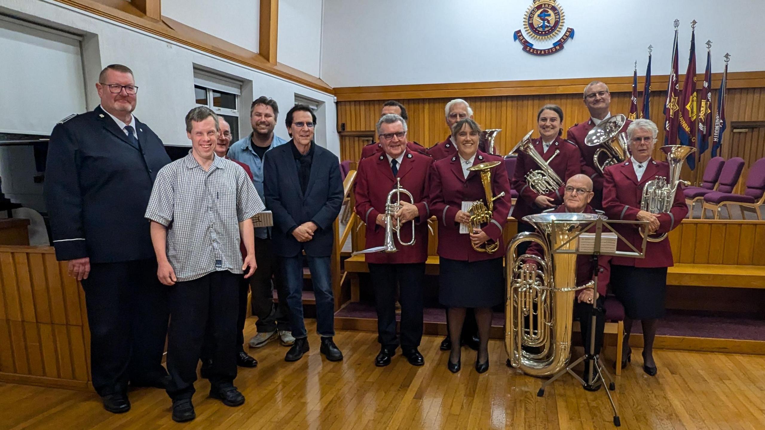 Shakin' Stevens stood in the centre wearing a blazer and some jeans next to people from the charity and the brass band who are wearing red uniforms.