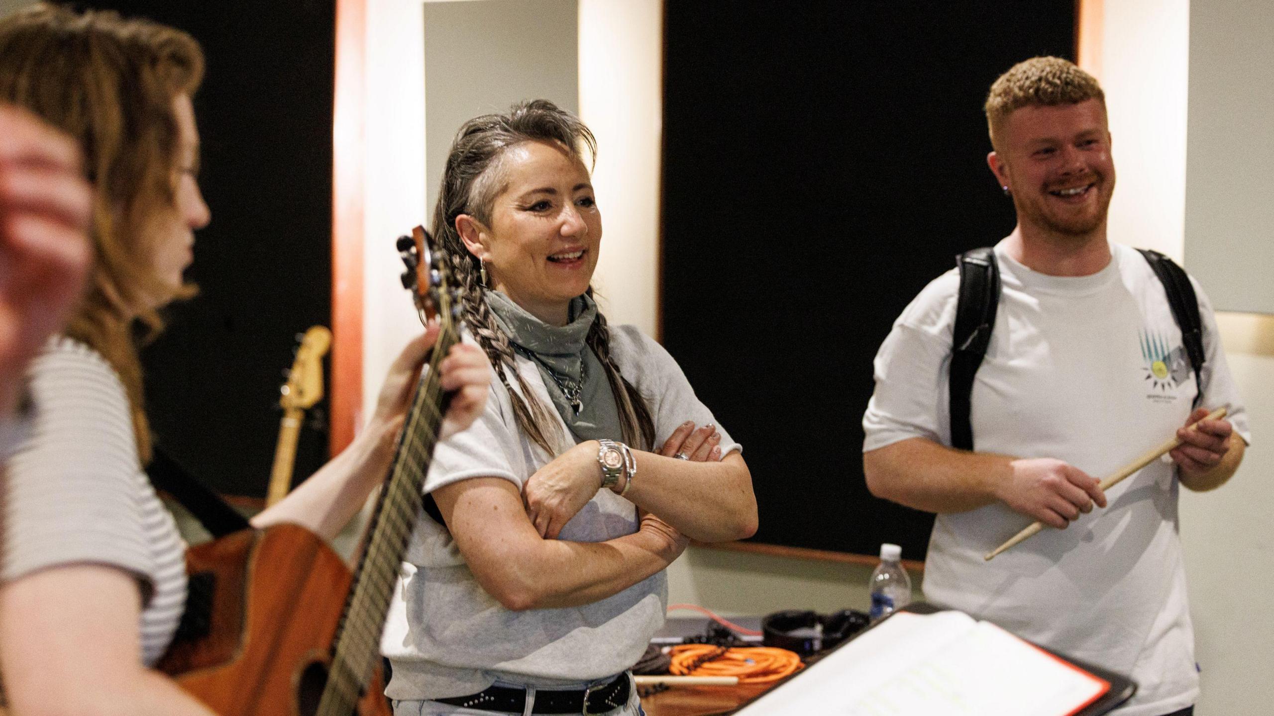 KT Tunstall in white T-shirt and scarves is standing in a bare rehearsal room with Jamie Rea, also in white T-shirt. They are surrounded by musicians.