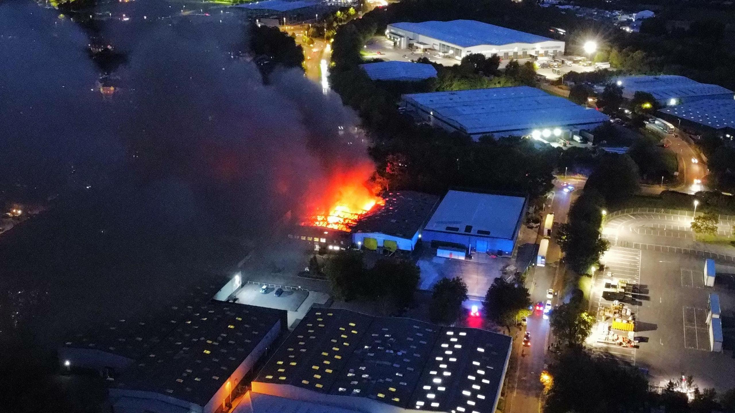 Drone footage shows a series of industrial units in the dark. In the middle is one unit on fire with red flames and smoke coming from the top.