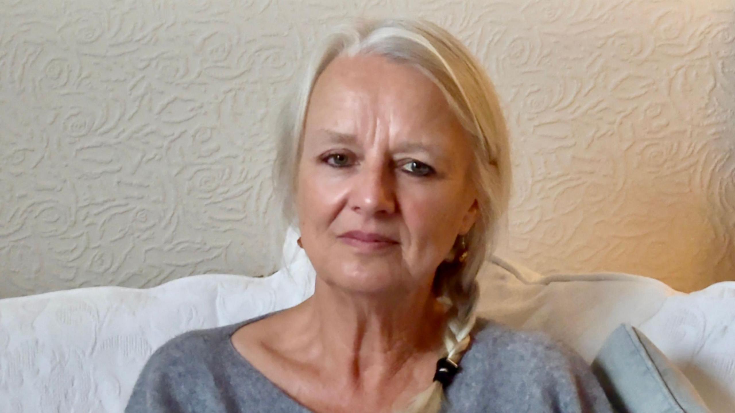 A close-up image of Sheila Tooth. She has long blond hair worn in a plait. She is wearing a grey top and is sitting on a white chair with a cream wall behind.