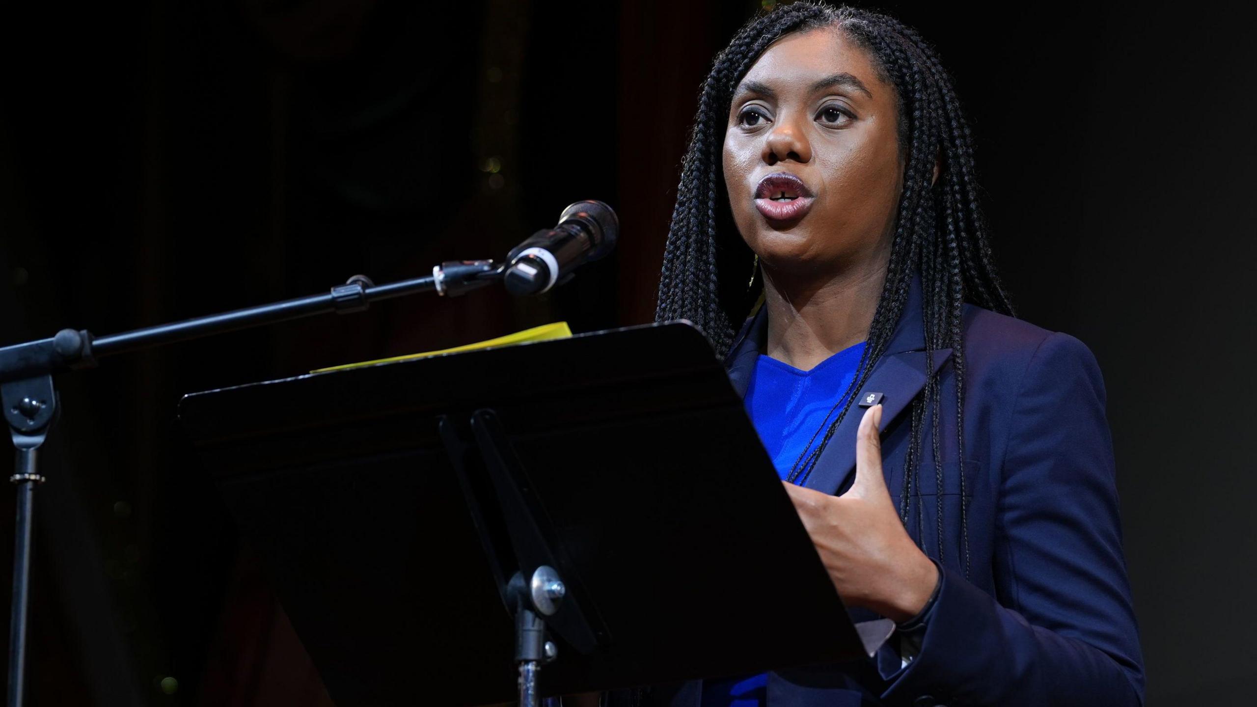 Conservative leader Kemi Badenoch, standing behind a black lectern on a stage, speaks to an audience  