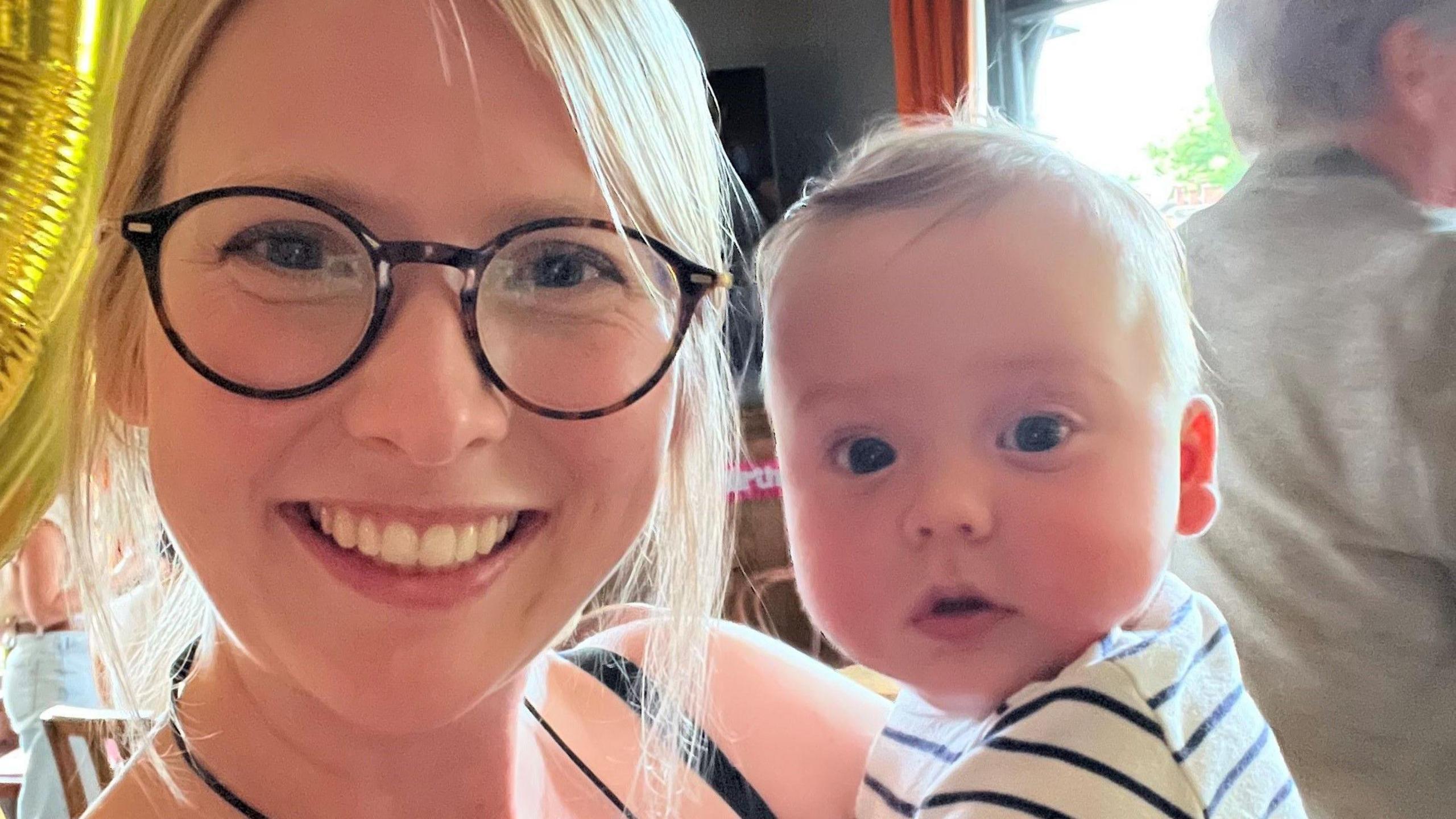 Andrea Gooch has blonde hair and is wearing glasses with tortoise shell rims. She is holding Austin, who is a baby wearing a white and black striped top.