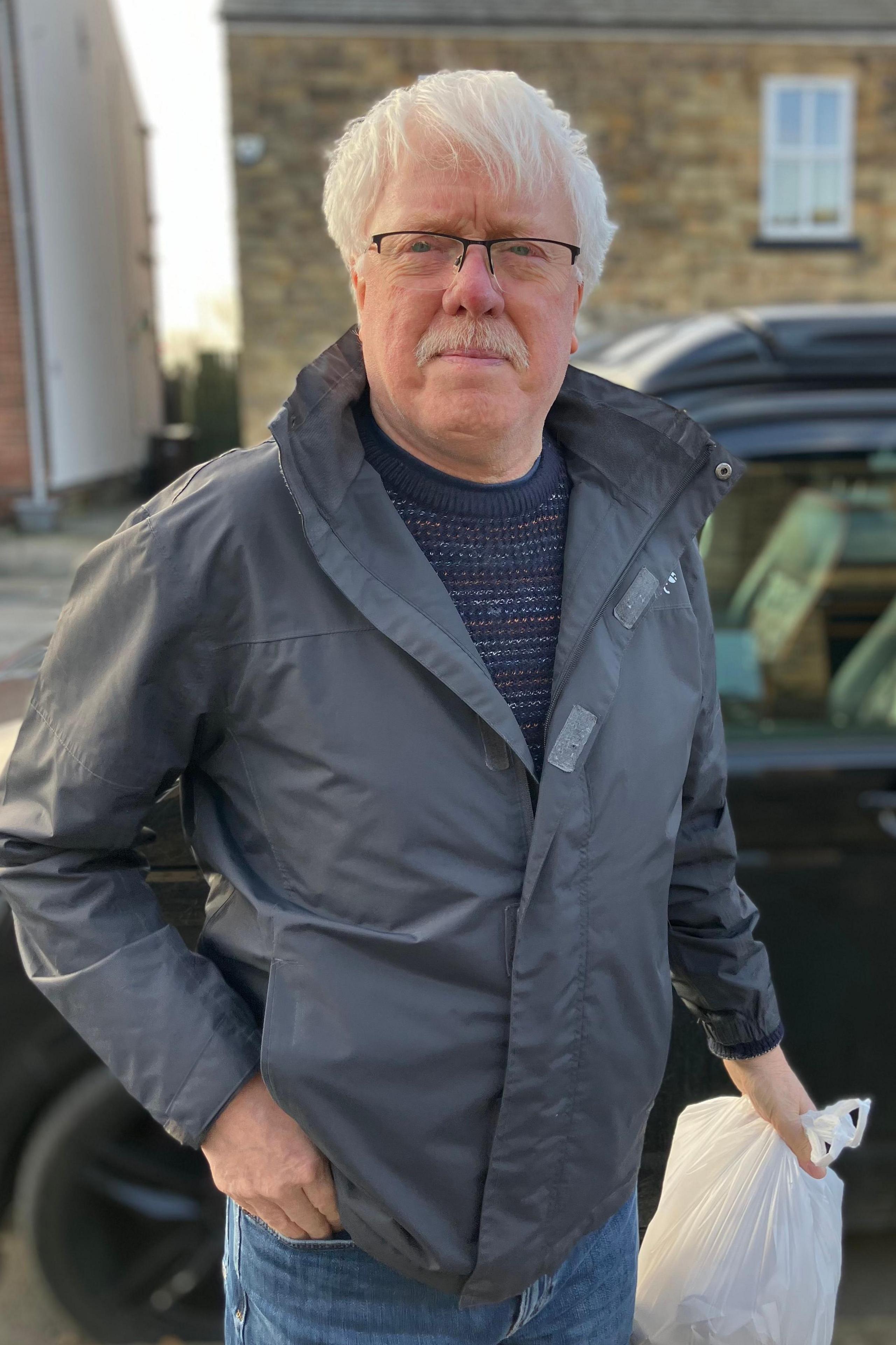 Paul Gilbert, who has white mid-length hair and a moustache. He is also wearing glasses and holding a white carrier bag.