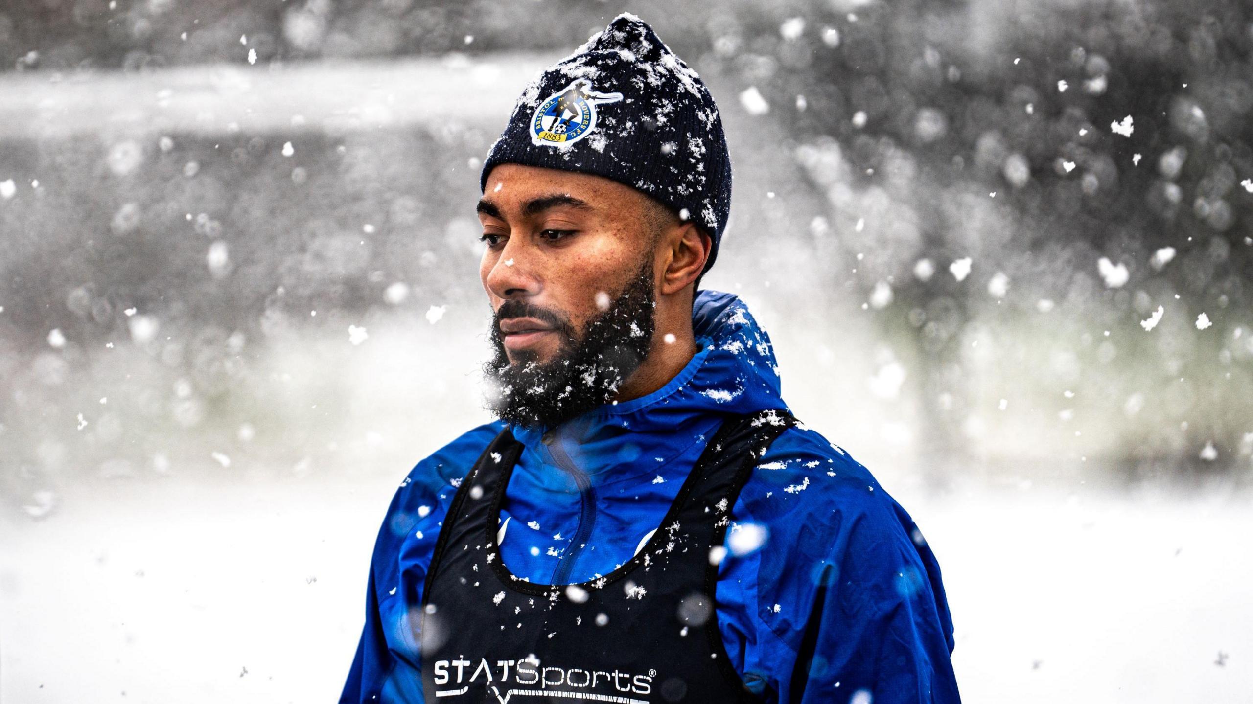 A Bristol Rovers player stands still during a training session with heavy snow falling. He has a blue jacket on with a black vest over the top of it, and also a black knitted hat with the Bristol Rovers club badge on it