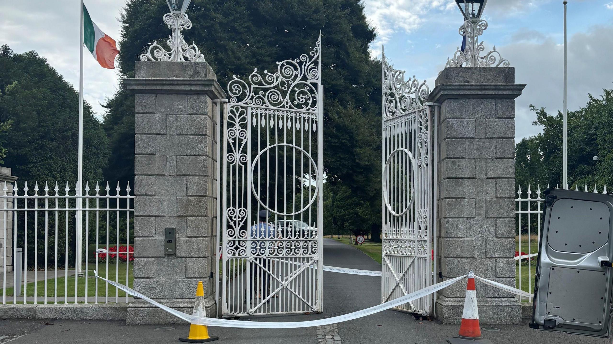 The gates at Dublin's Phoenix Park, the official residence of Ireland's President, Michael D Higgins. The white gates are pictured behind police cones and tape and are partially open.