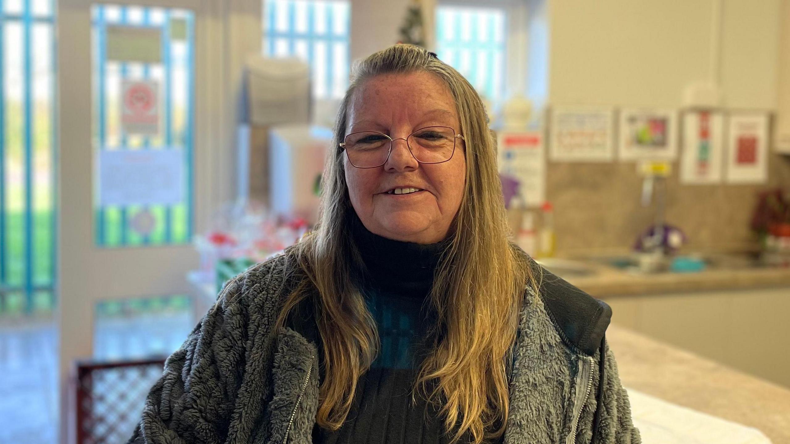 Sally Grieff is wearing a fleece and turtleneck, she is standing in front of the counter where they exchange money and food boxes.