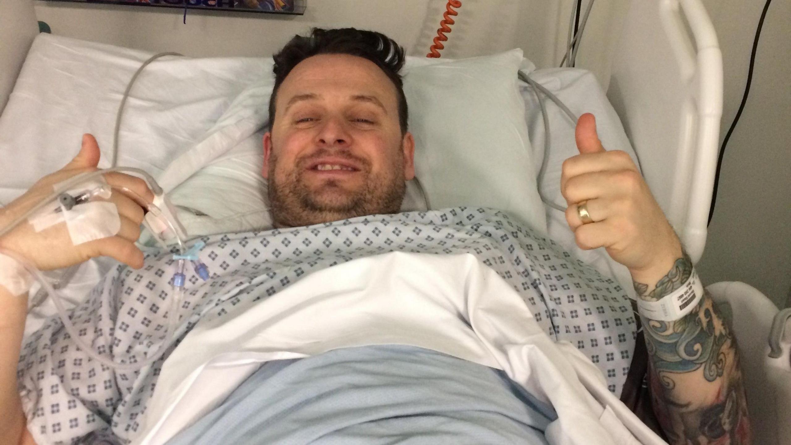 Vince in a hospital gown in a hospital bed with tubes going into one hand and he's is giving a double thumbs up and smiling