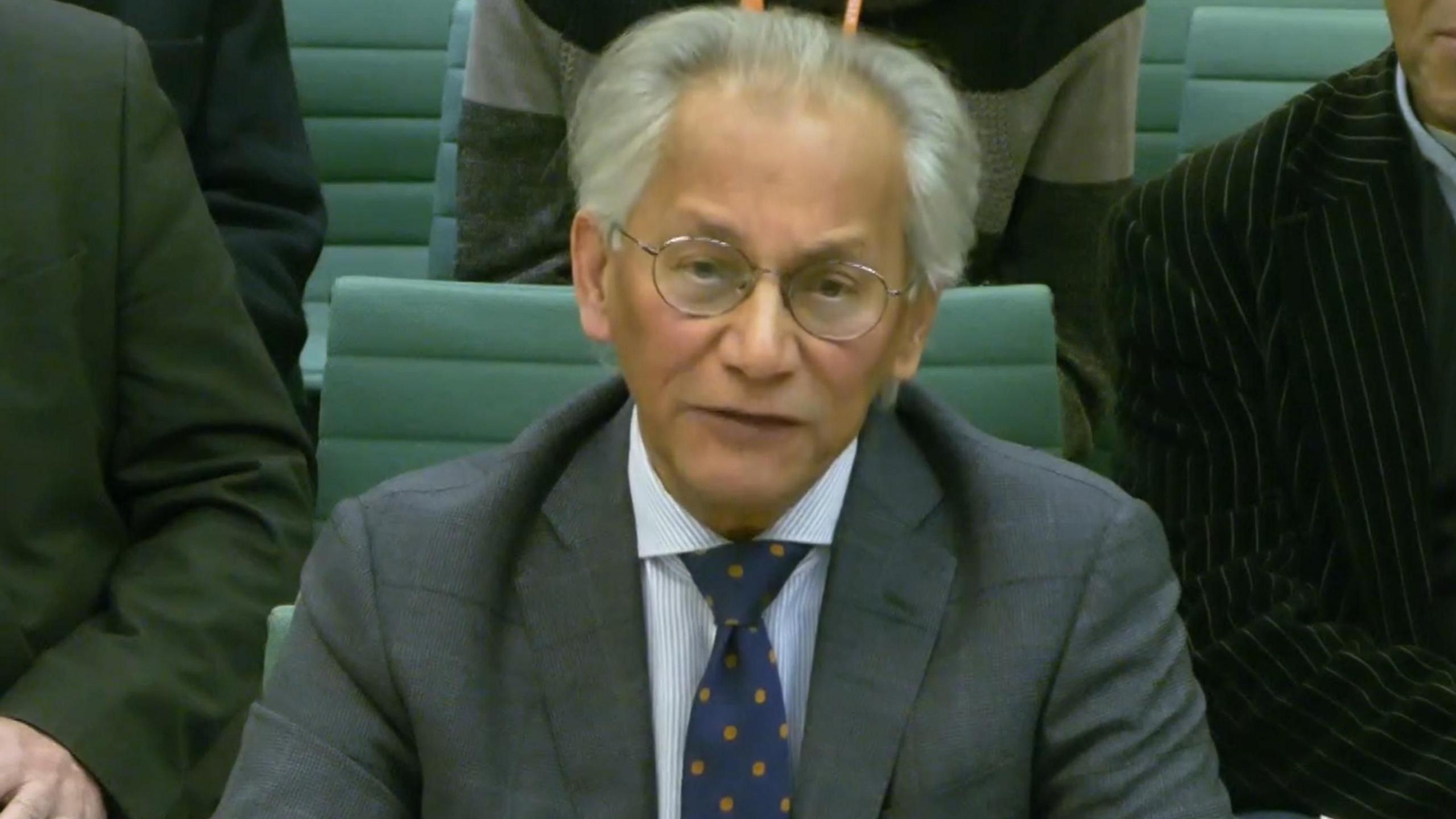 Samir Shah giving evidence at the committee - he is sitting on a green chair, with other people behind him. He has grey hair and thin-rimmed glasses and is wearing a grey suit jacket with a striped shirt and blue spotted tie