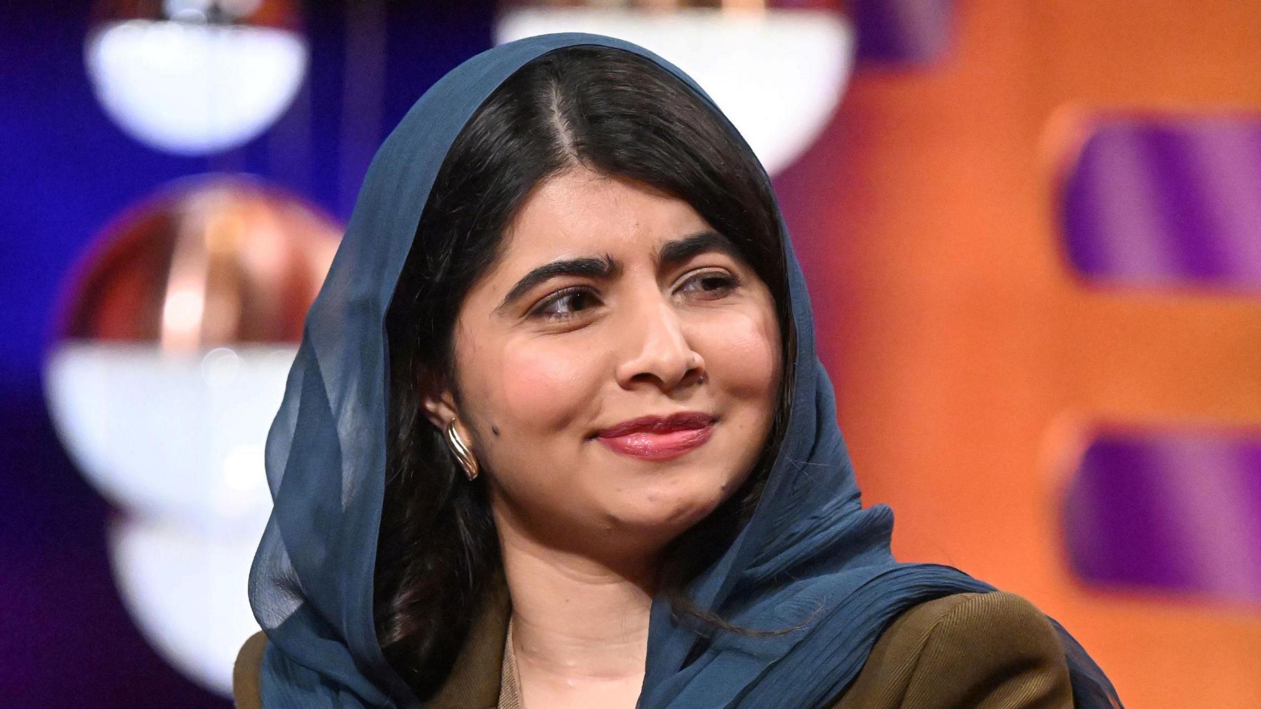 Malala Yousafzai looks to the side smiling. She has long dark hair and is wearing a blue head scarf and dark jacket.
