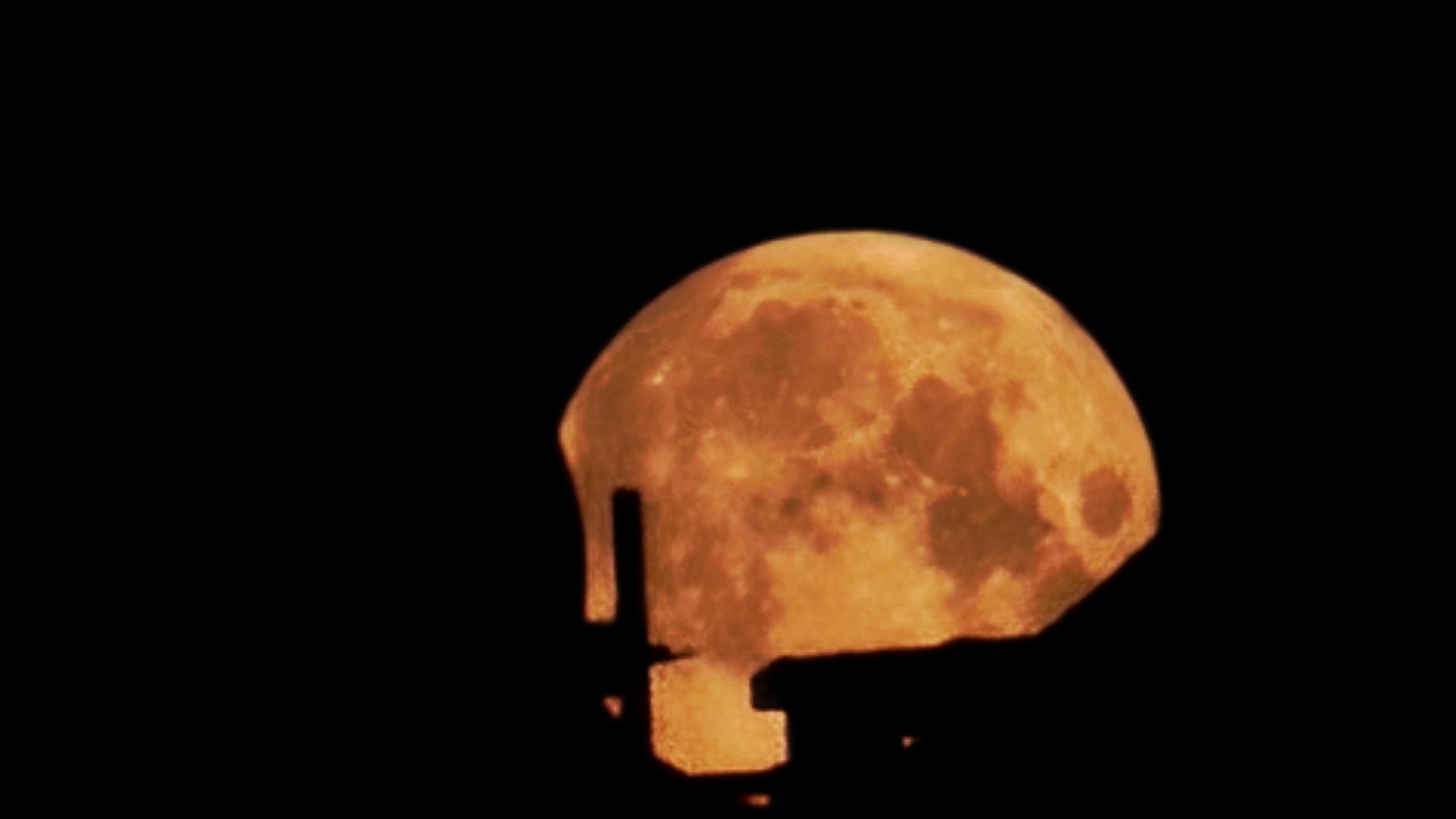 Orange coloured full Moon peaks out behind silhouetted buildings