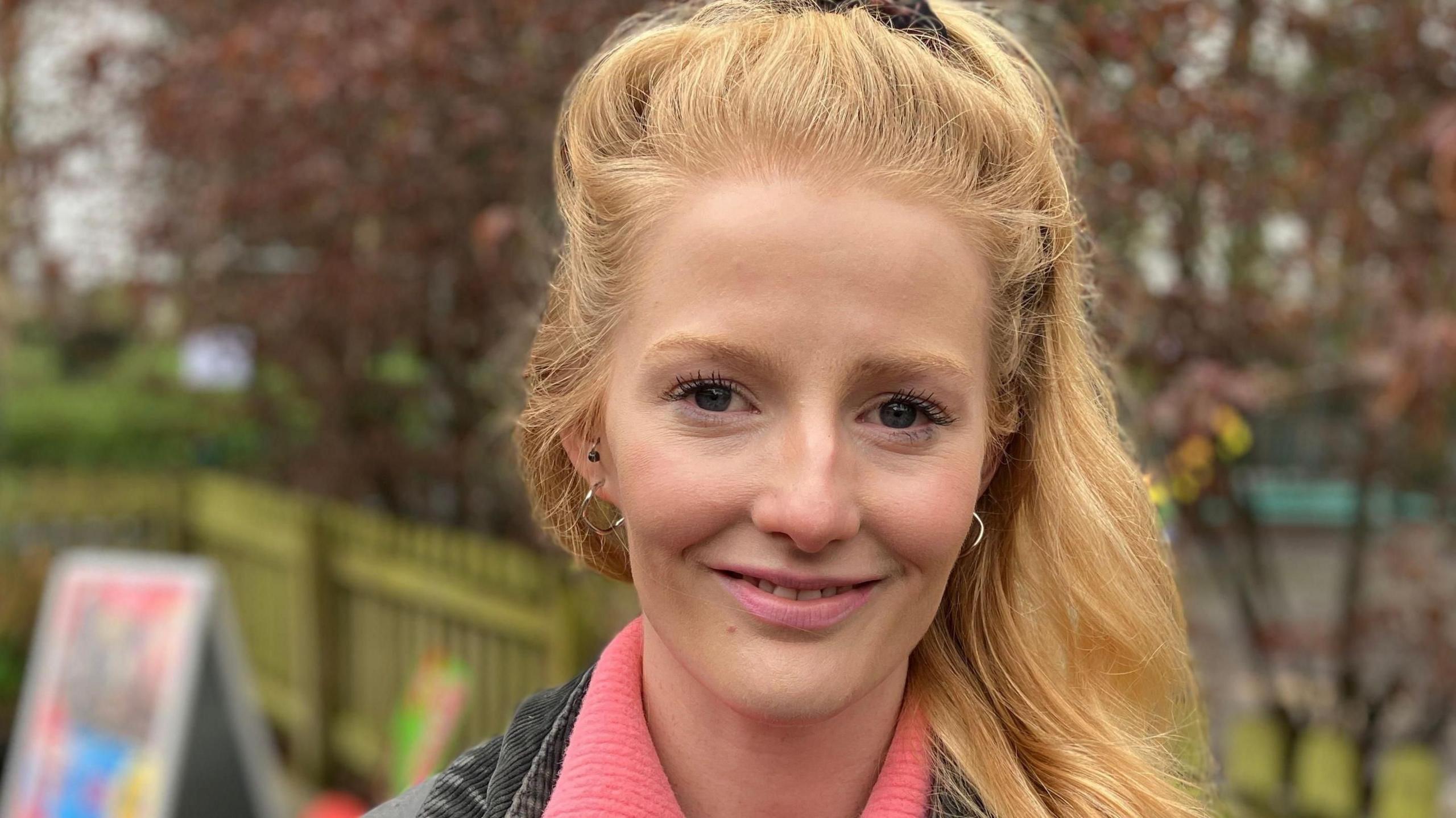 Hannah Spencer has long blonde hair and is wearing a black denim jacket and pink sweater. She is standing outdoors in a playground.