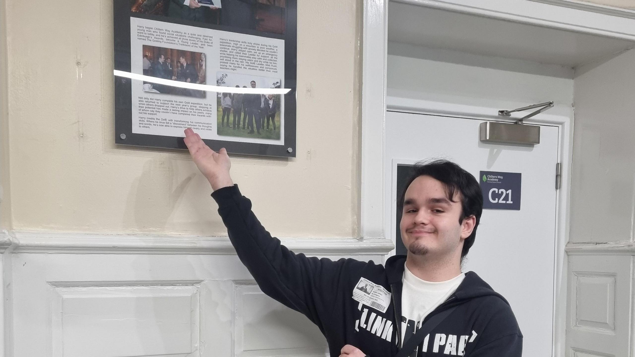 Harry pointing at a picture of his Duke of Edinburgh award in a hallway, he is smiling and looking at the camera, wearing a black hoodie, and white top. He is by a white door, and walls with panelling on. 