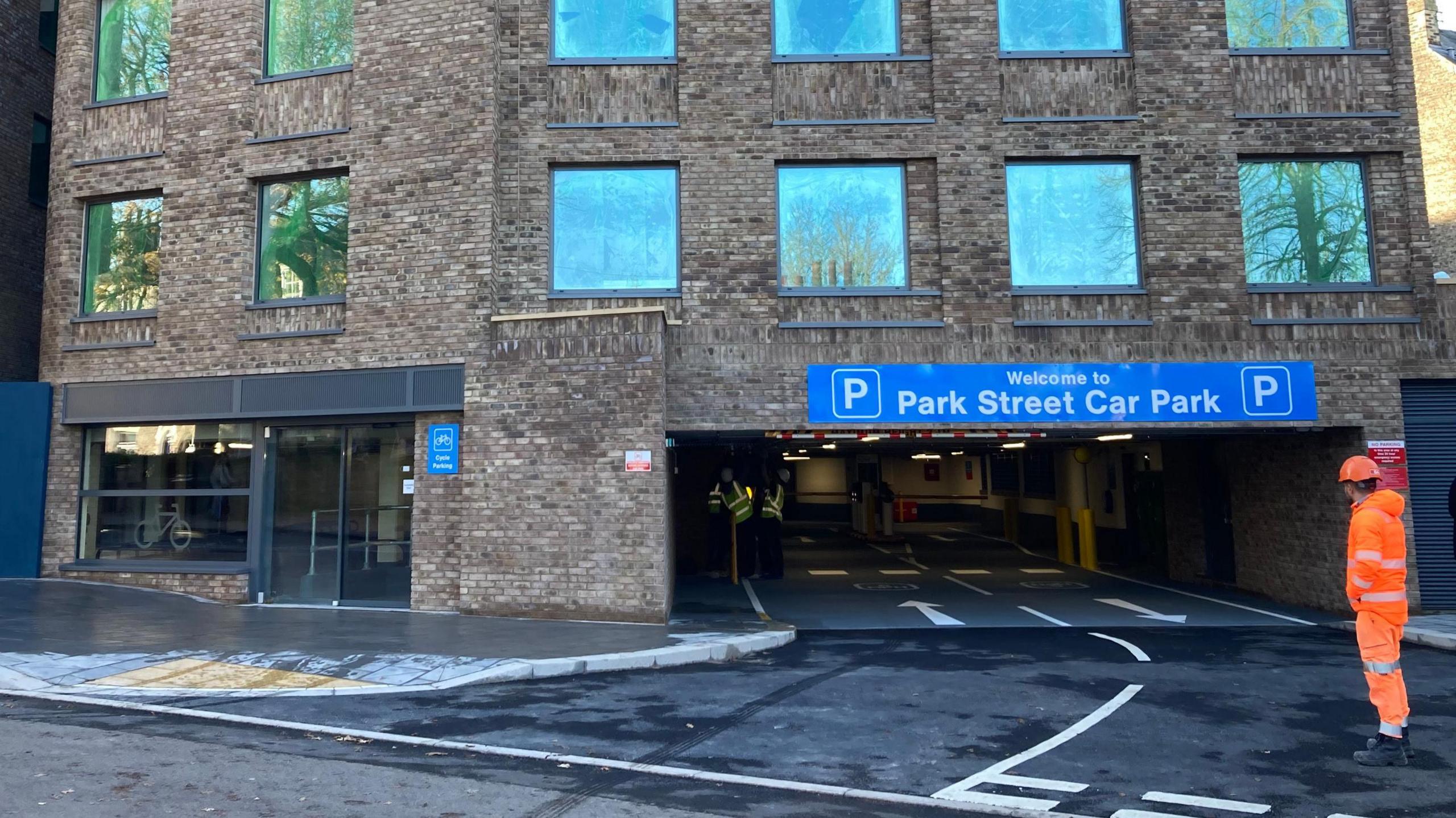 The Park Street car park. A man in an orange fluorescent outfit is stood outside the front of the entrance in a hard hat. A blue sign on the entrance and exit of the car park says, Welcome to Park Street Car Park in white letters.