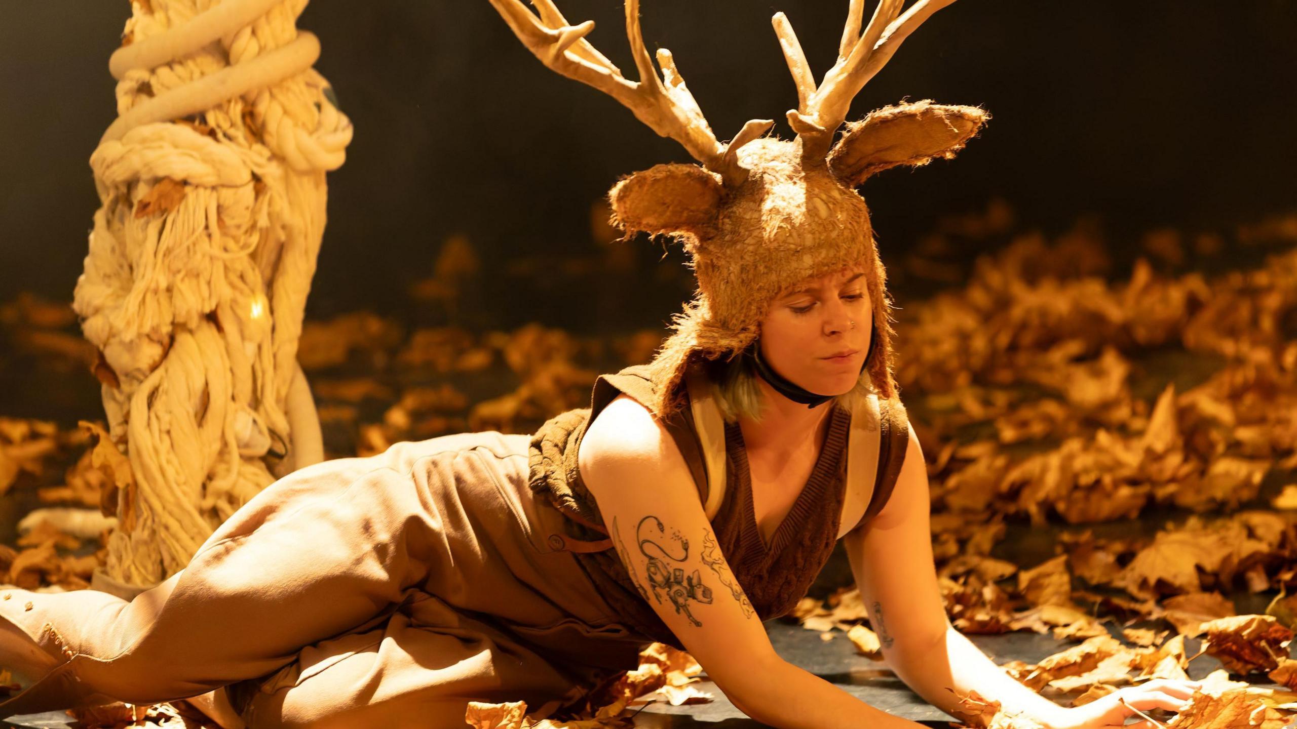 A woman rehearses for a Circomedia show called Mother Deer in Bristol. She has a fur-type headdress on, with antlers attached, and is lying down on a bed of leaves