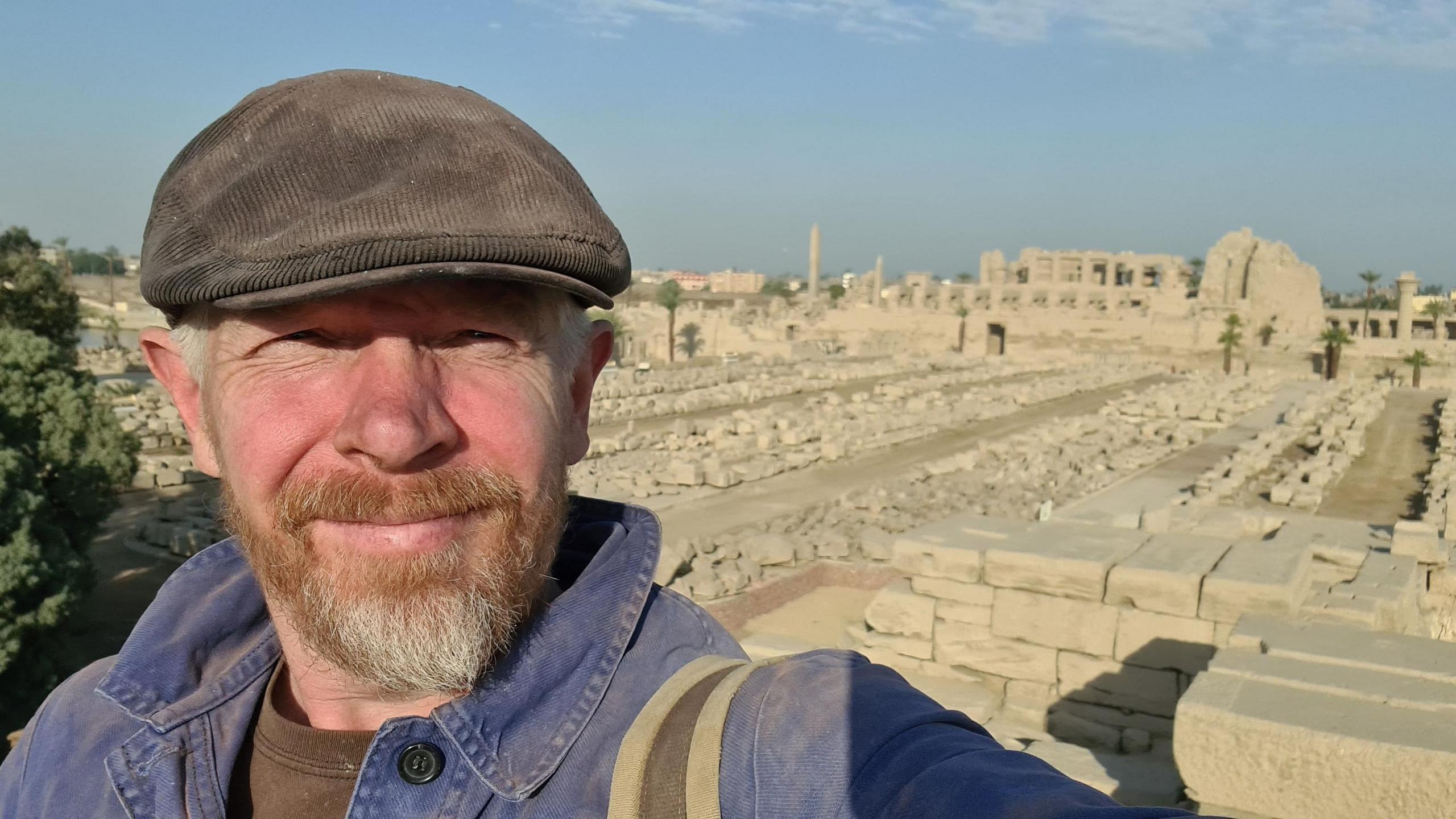 Theo Gay Anderson in flat cap and blue denim shirt has a brown greying beard and grey hair.