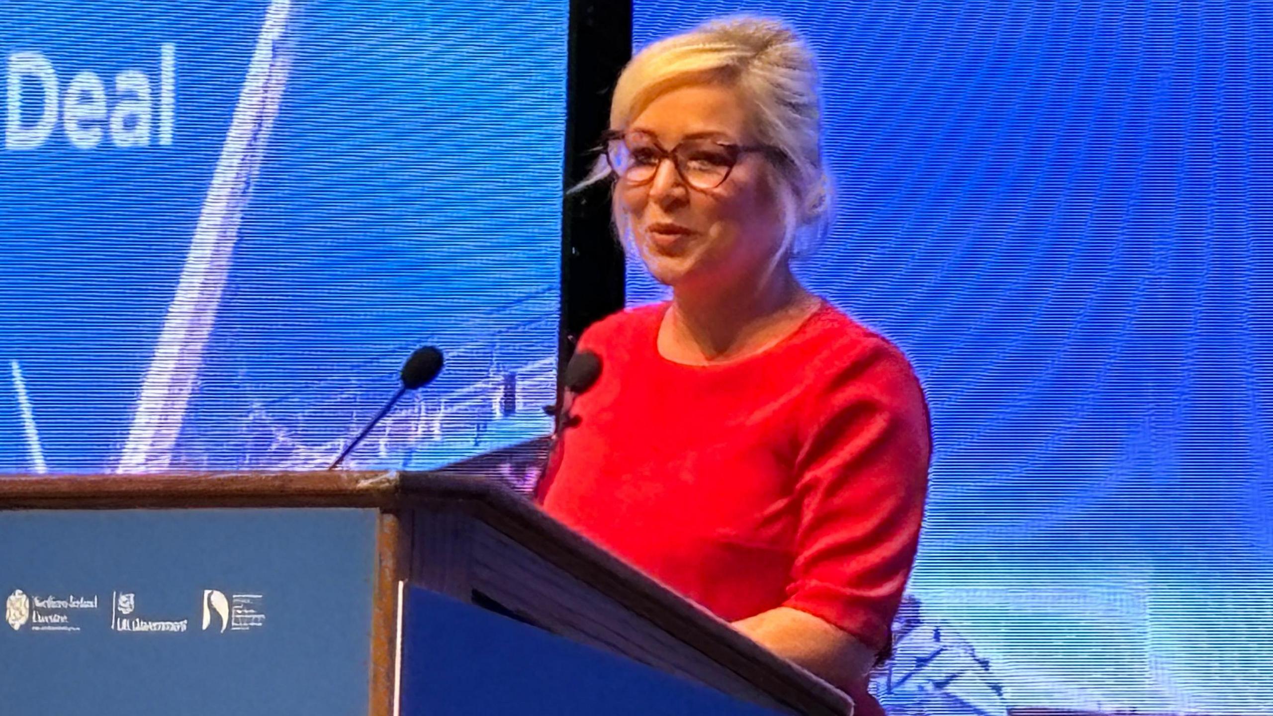 Shows Michelle O'Neill in a red dress at a microphone 