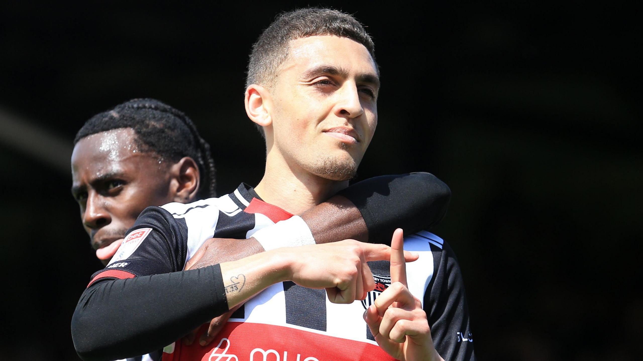 Jaze kabia celebrates a goal by holding his fingers in a T-shape in front of him whilst being hugged round neck by teammate, wearing Grimsby black and white striped home kit