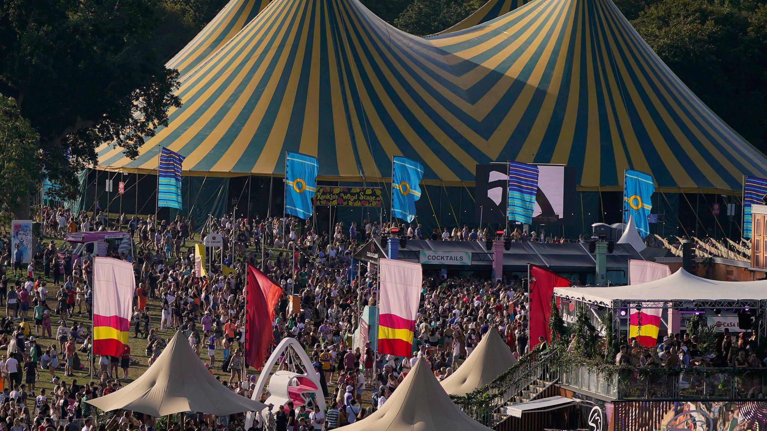 Two large yellow and blue tents with hundreds of people stood below them, numerous colourful flags are billowing and there are several sound stages