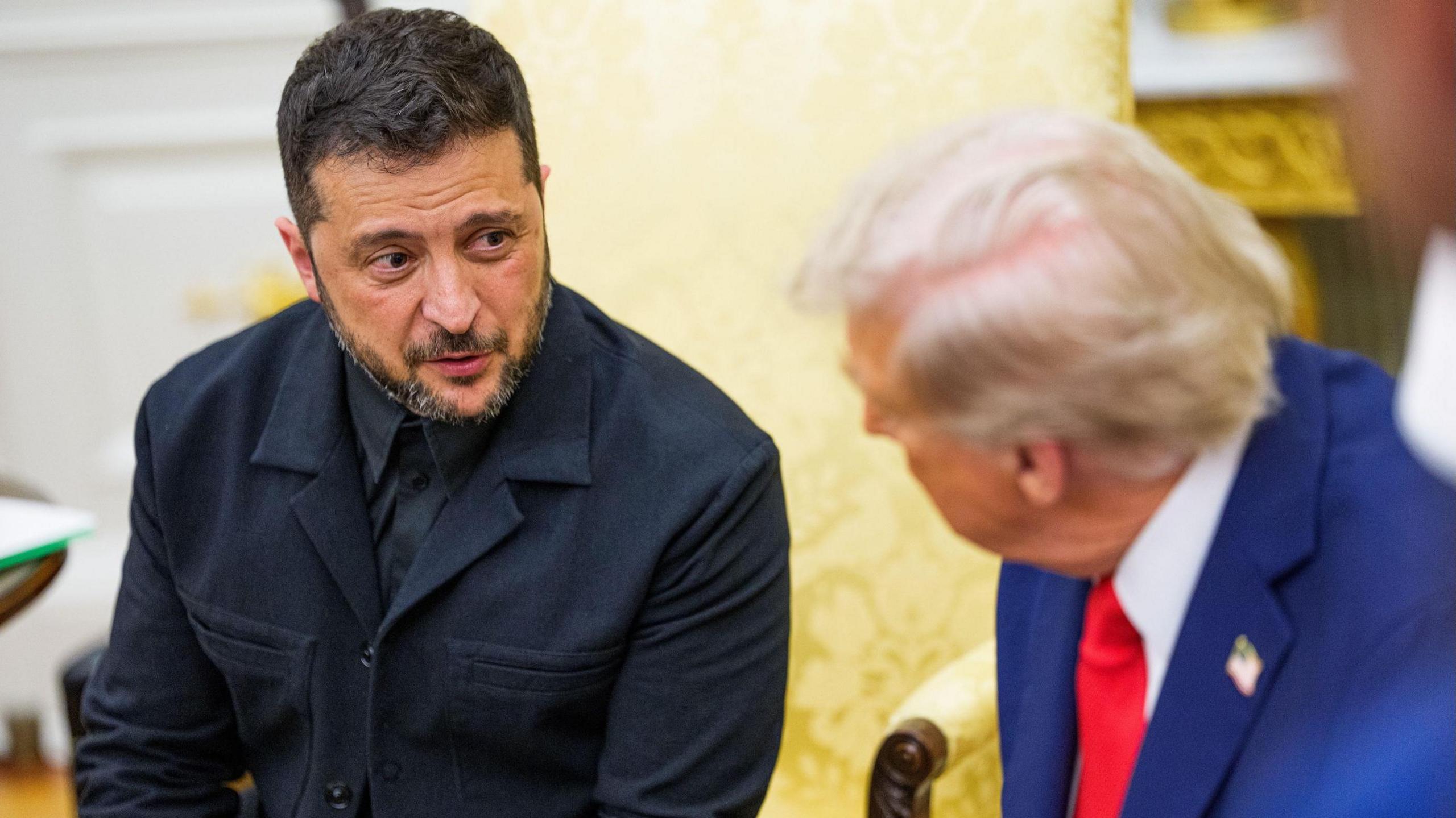 Ukrainian President Zelensky wears a dark suit and looks towards US President Donald Trump, in a blue suit with red tie.