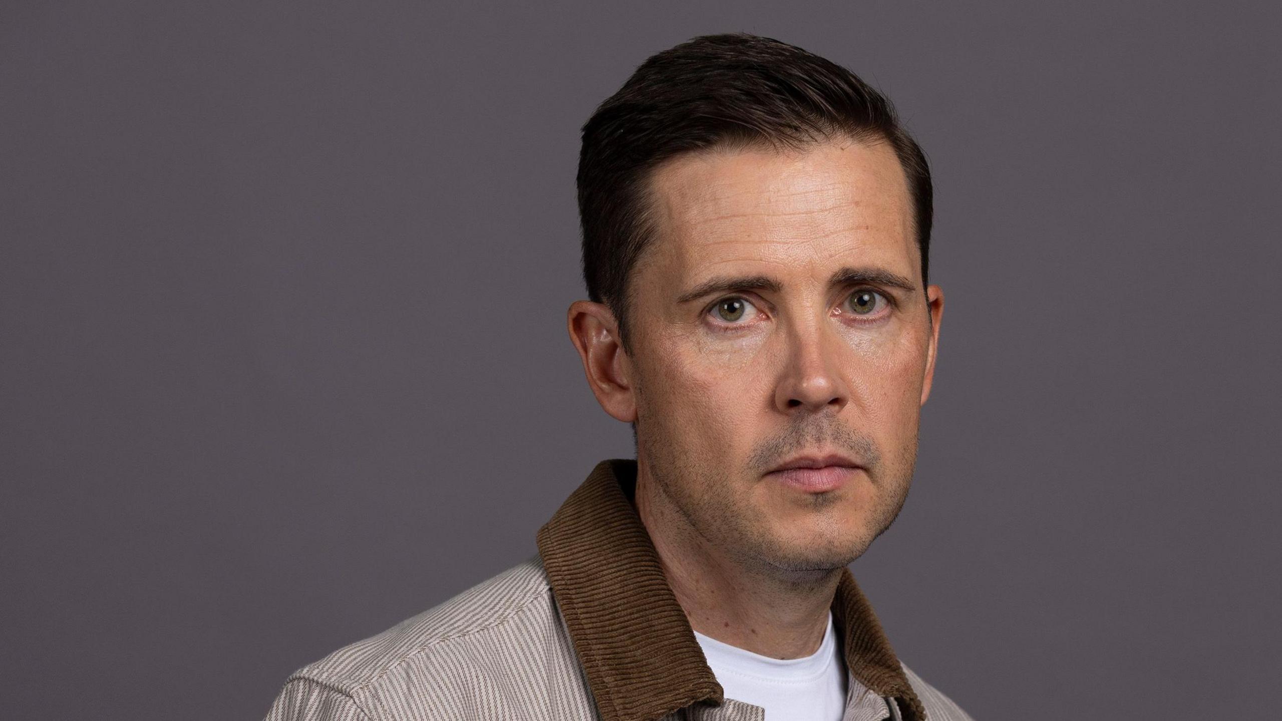 Gareth Pierce, who plays the part of Todd Grimshaw in Coronation Street, has short dark hair and hazel eyes. He is clean shaven and wears a brown-and-white striped shirt over a white t-shirt.