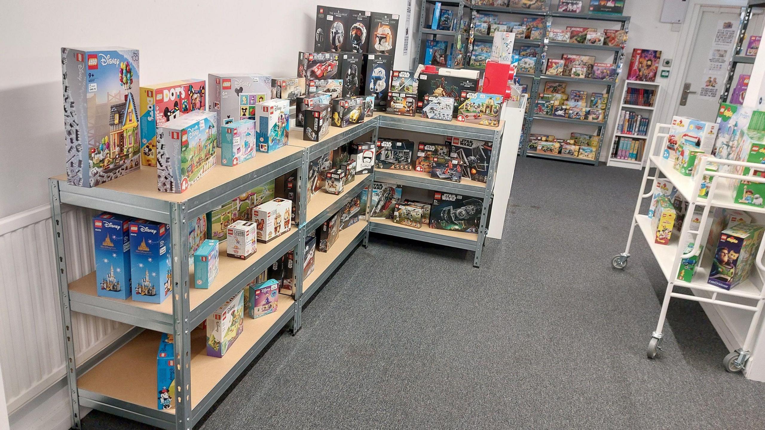 Metal shelves covered in Lego sets in a white room with grey carpet.